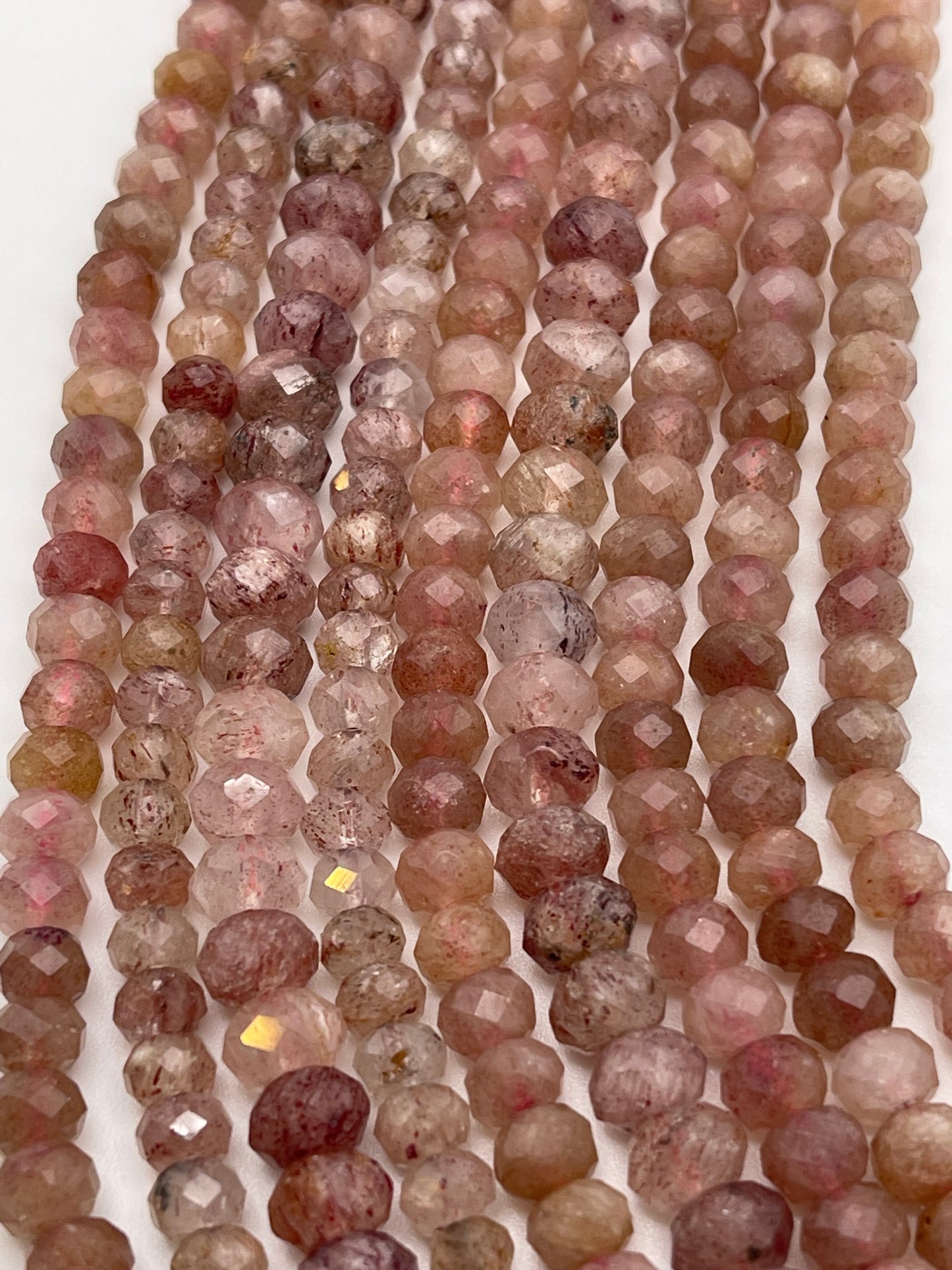 Close-up image of multiple strands of 6mm Fac Strawberry Quartz Rondell beads in varying shades of pink, brown, and beige. The beads have a translucent quality and some exhibit specks of other colors, creating a textured and varied appearance.