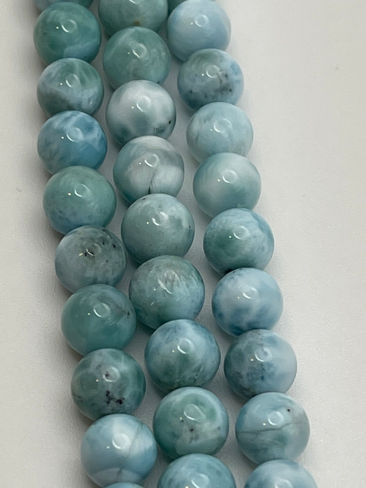 Close-up image showing strands of 8mm Larimar Rnd Smooth beads in varying shades of blue and green, arranged neatly on a white background. The beads have a glossy finish with subtle color variations and natural patterns.
