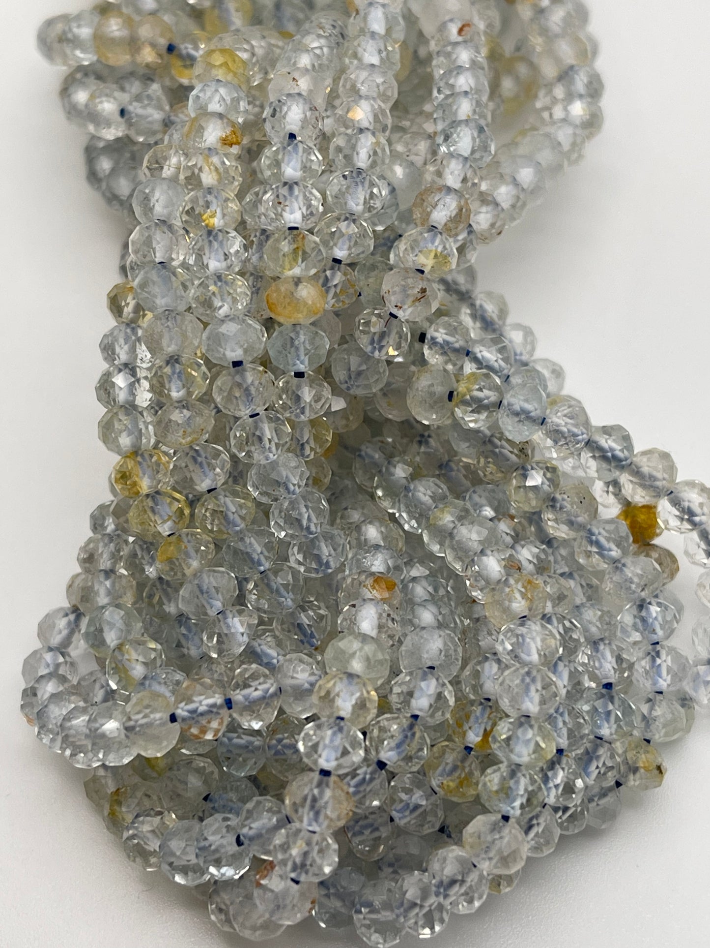 A close-up of a bundle of 5mm Fac Topaz Rondell beads with a mix of clear, yellow, and blue hues. The beads are intricately strung together, creating a sparkling, textured appearance against a solid white background.