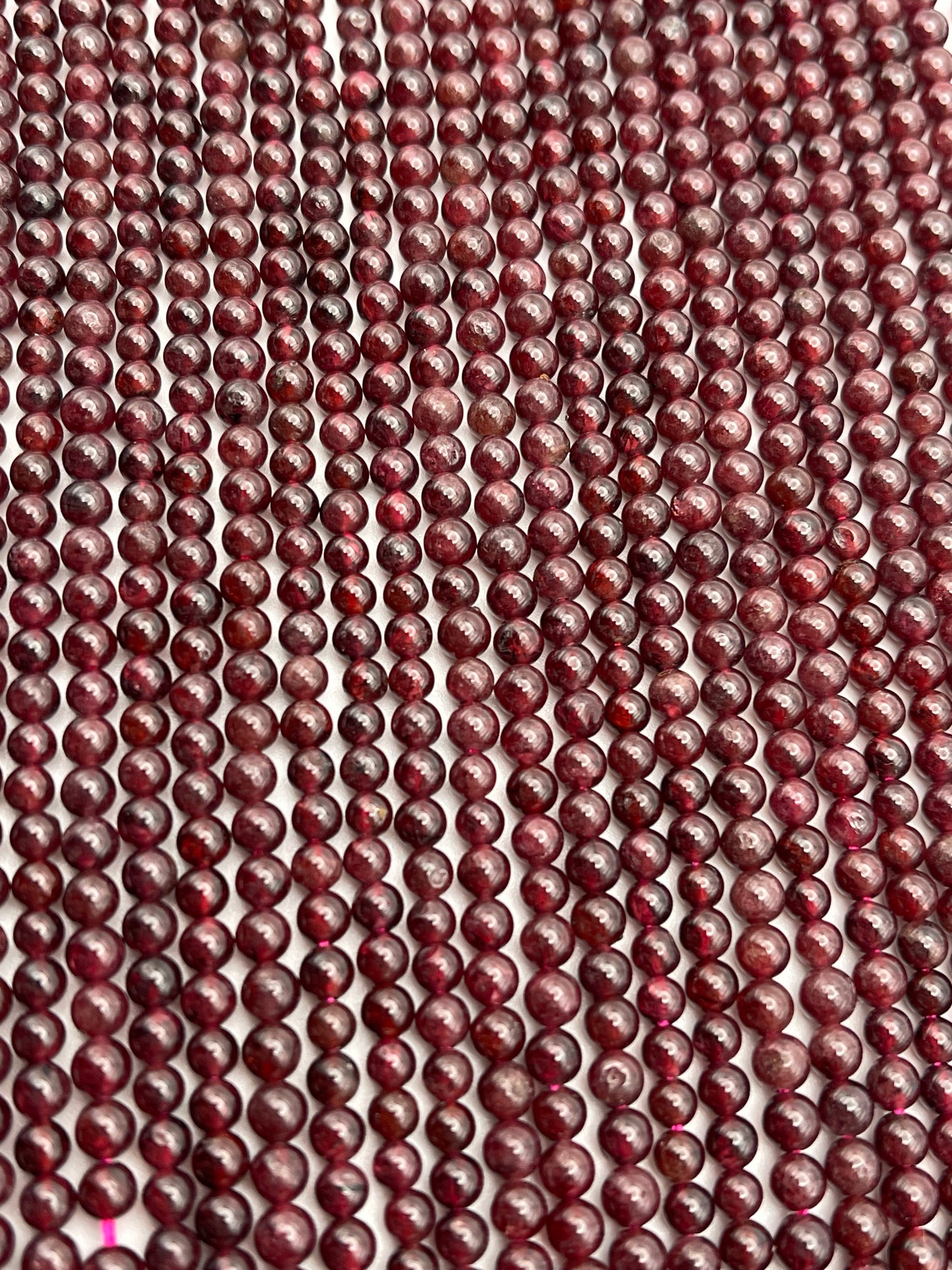 Close-up view of numerous 4mm Round Garnet beads aligned in parallel rows, creating a textured and pattern-rich visual. The surface of each garnet bead exhibits slight variations in color and sheen, adding to the intricate detail of the arrangement.
