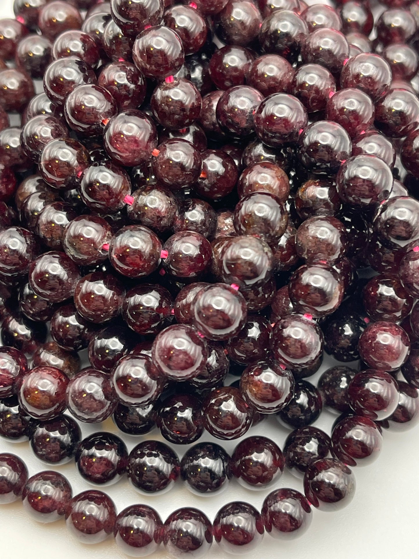 Close-up of several strands of 8mm Garnet Rnd Smooth beads. The spherical, dark red garnet beads are tightly packed and display a glossy surface that reflects light, giving a rich, lustrous appearance. These uniformly sized garnet beads are strung in continuous loops.