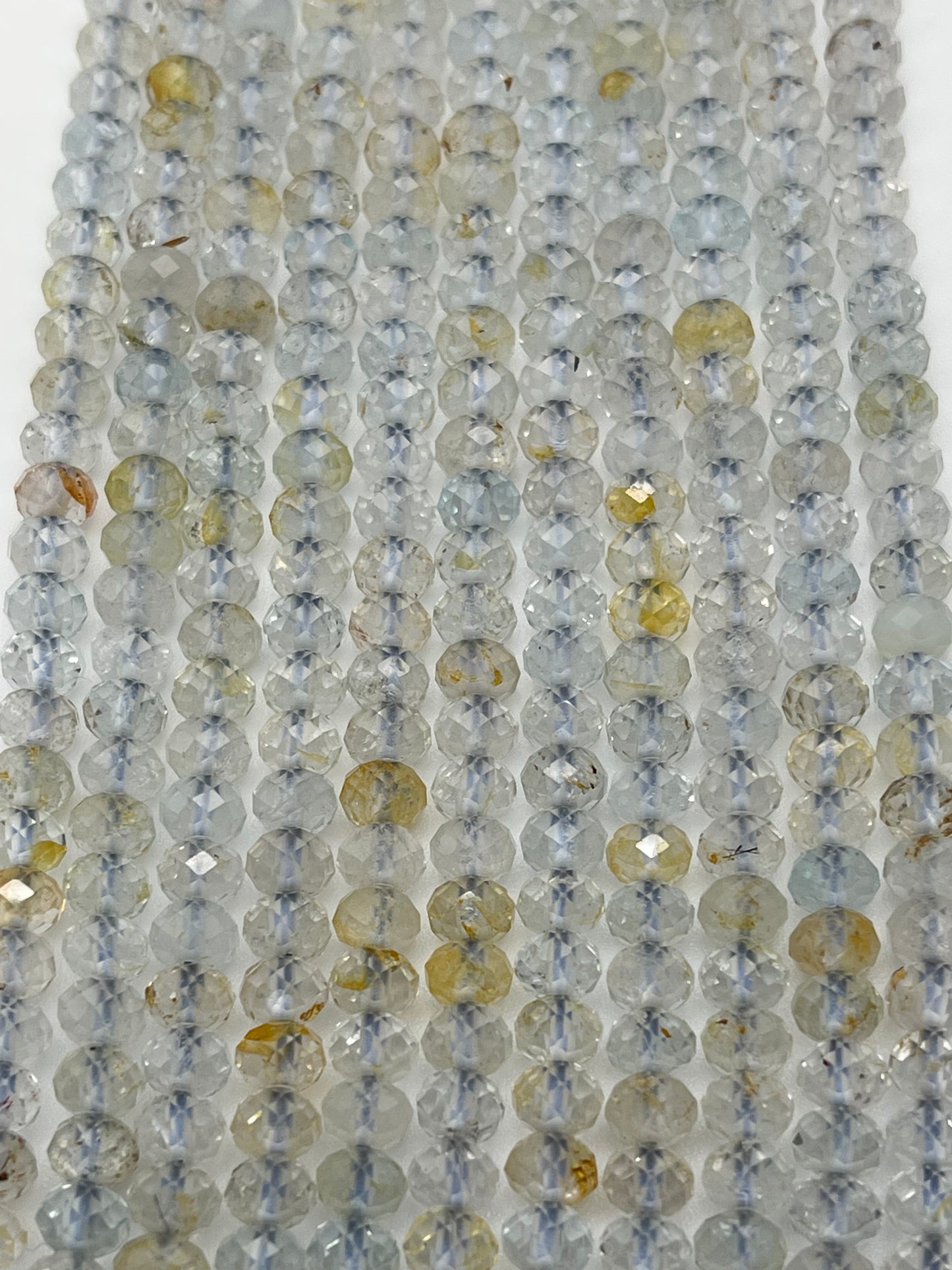 Strings of 5mm Fac Topaz Rondell beads in an array of light gray and yellow hues are strung closely together. The beads have a translucent quality with some displaying flecks of brown and other colors, creating a shimmering effect under the light.
