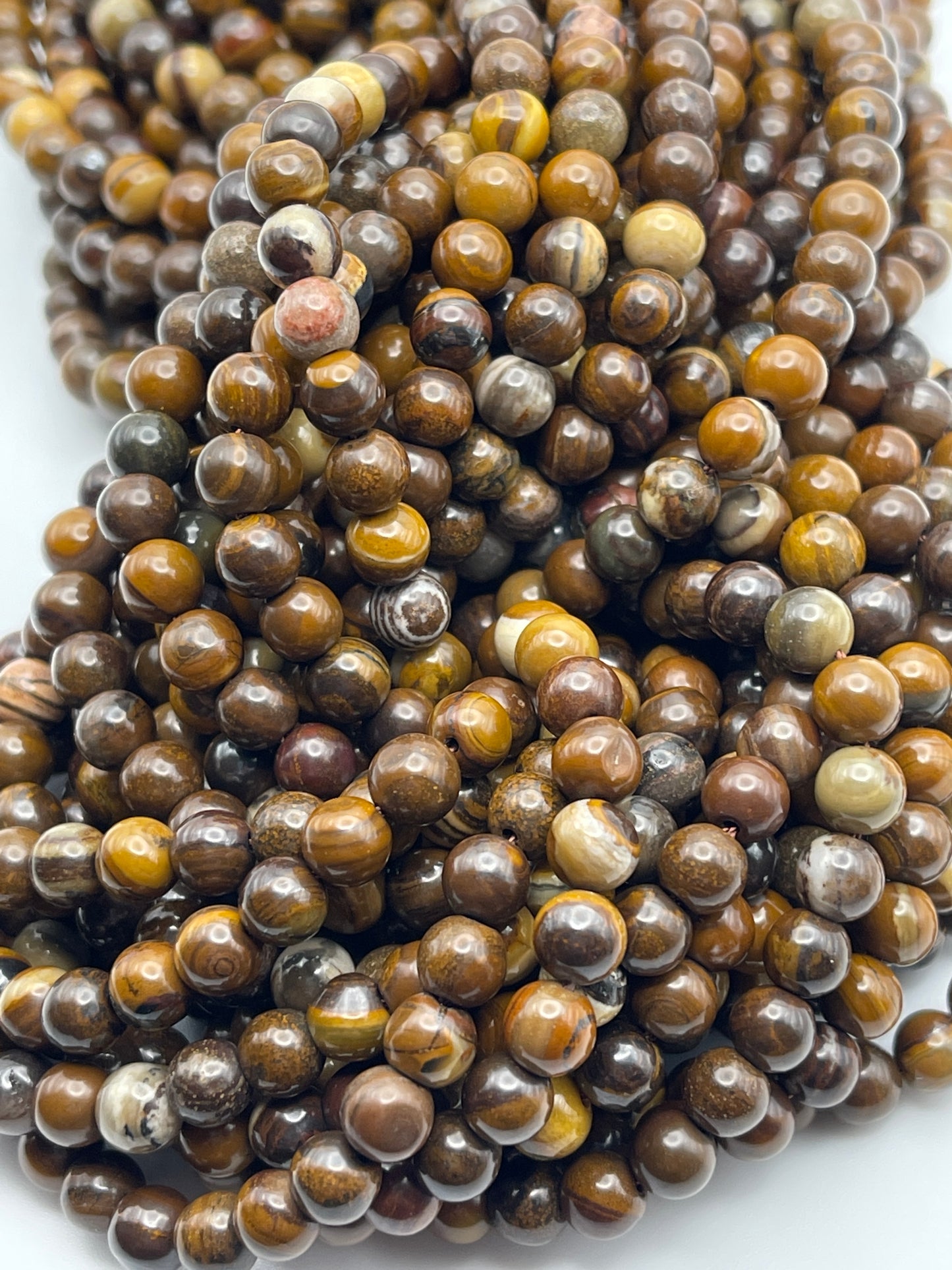 A heap of polished 6mm Tiger Iron Round Jasper beads, intricately strung together in varying shades of brown and tan. The beads exhibit rich, marbled patterns, creating a versatile and earthy visual texture.