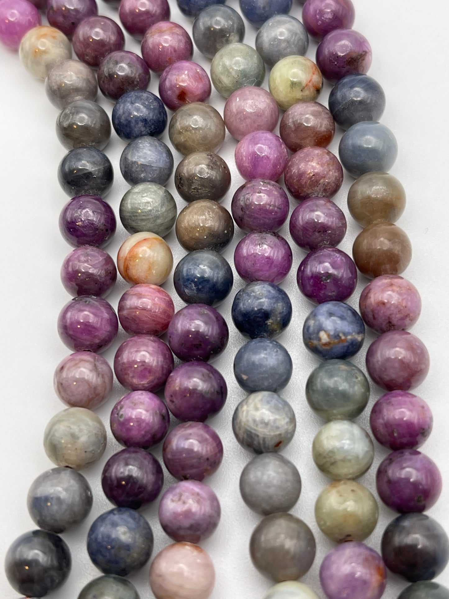 A close-up of several strands of 8mm Sapphire Rainbow Rnd Smooth beads in various shades of purple, pink, blue, gray, and green. Each bead is unique in color, giving the strands a multicolored and natural appearance. The beads are placed closely together on a white background.