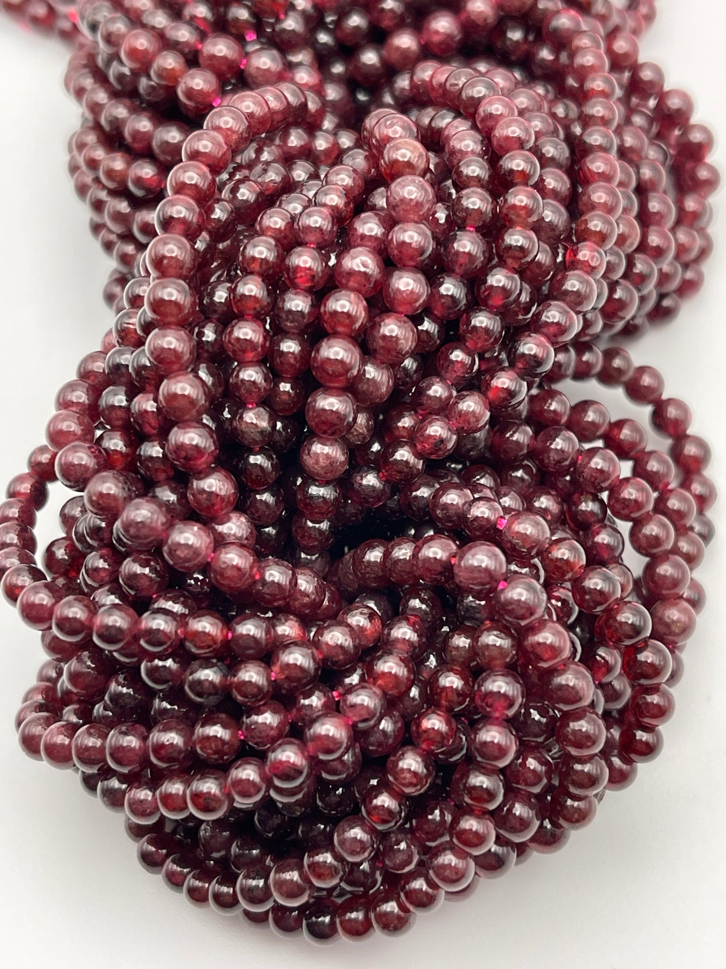 A close-up image of multiple strands of 4mm Round Garnet beads, tightly bundled together. The garnet beads are shiny, spherical, and uniform in size, creating a rich, glossy texture.