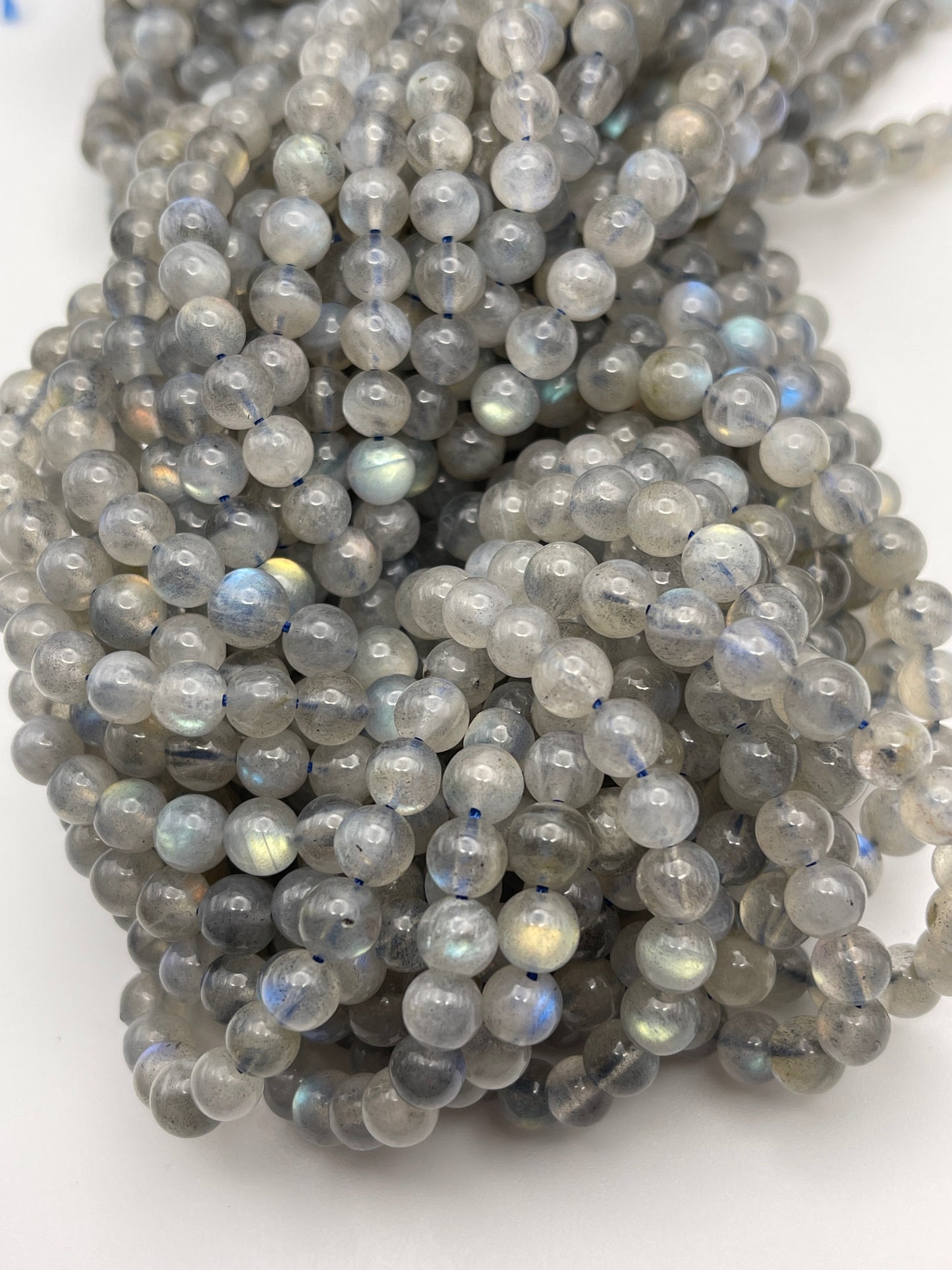 A close-up view of numerous 6mm Round Labradorite beads. The beads are predominantly grey with iridescent flashes of blue, green, and yellow. They are polished and arranged closely together, highlighting their smooth texture and semi-translucent appearance.