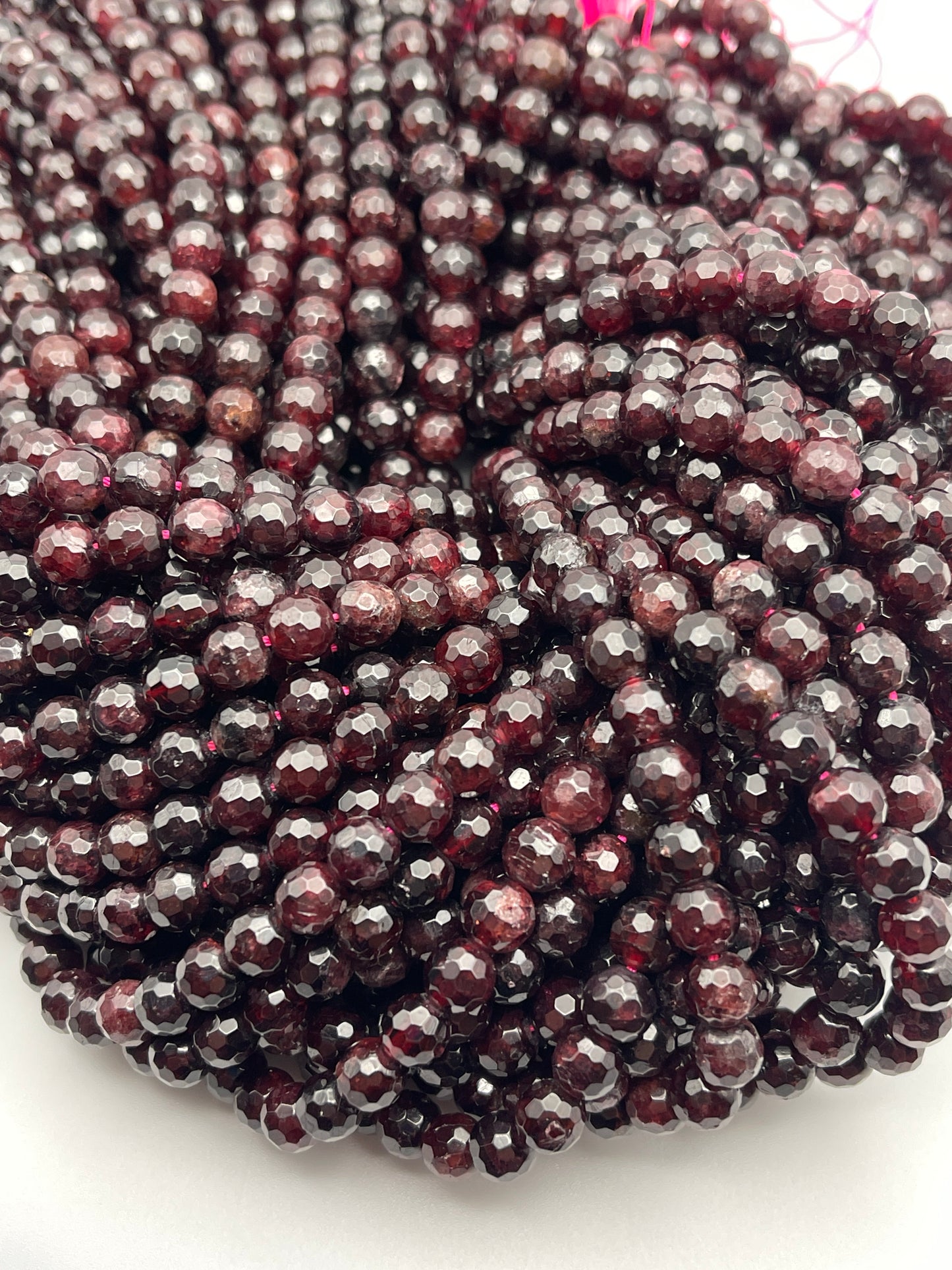 A close-up image of multiple strands of 7mm Faceted Garnet beads. The deep red, faceted beads are tightly clustered together, displaying a glossy sheen. The light reflections highlight the facets, adding to the richness and texture of the arrangement.