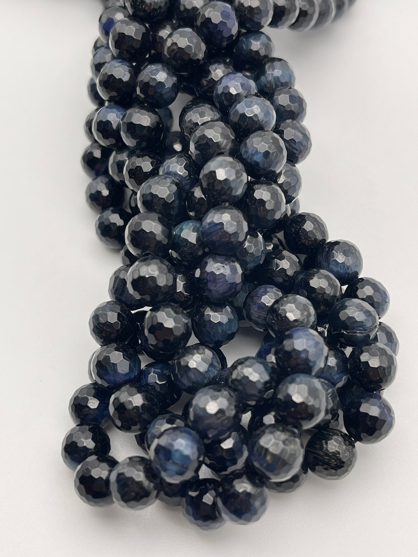 Close-up of several strands of polished 8mm Round Faceted Blue Tiger's Eye beads with a shiny and reflective appearance. The beads are tightly strung together, showcasing subtle variations in color and luster.