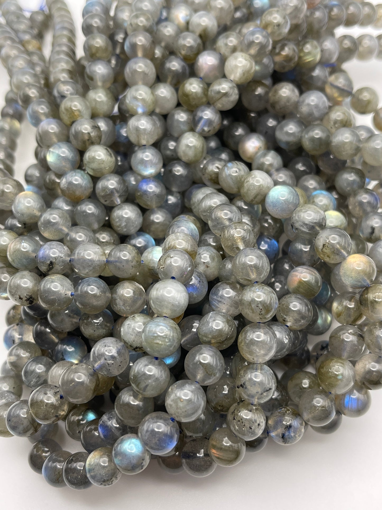 A collection of 8mm Labradorite Rnd Smooth PG beads in hues of gray and blue with iridescent reflections, producing a shimmering effect. The smooth, spherical beads are neatly organized in strands against a plain white background.