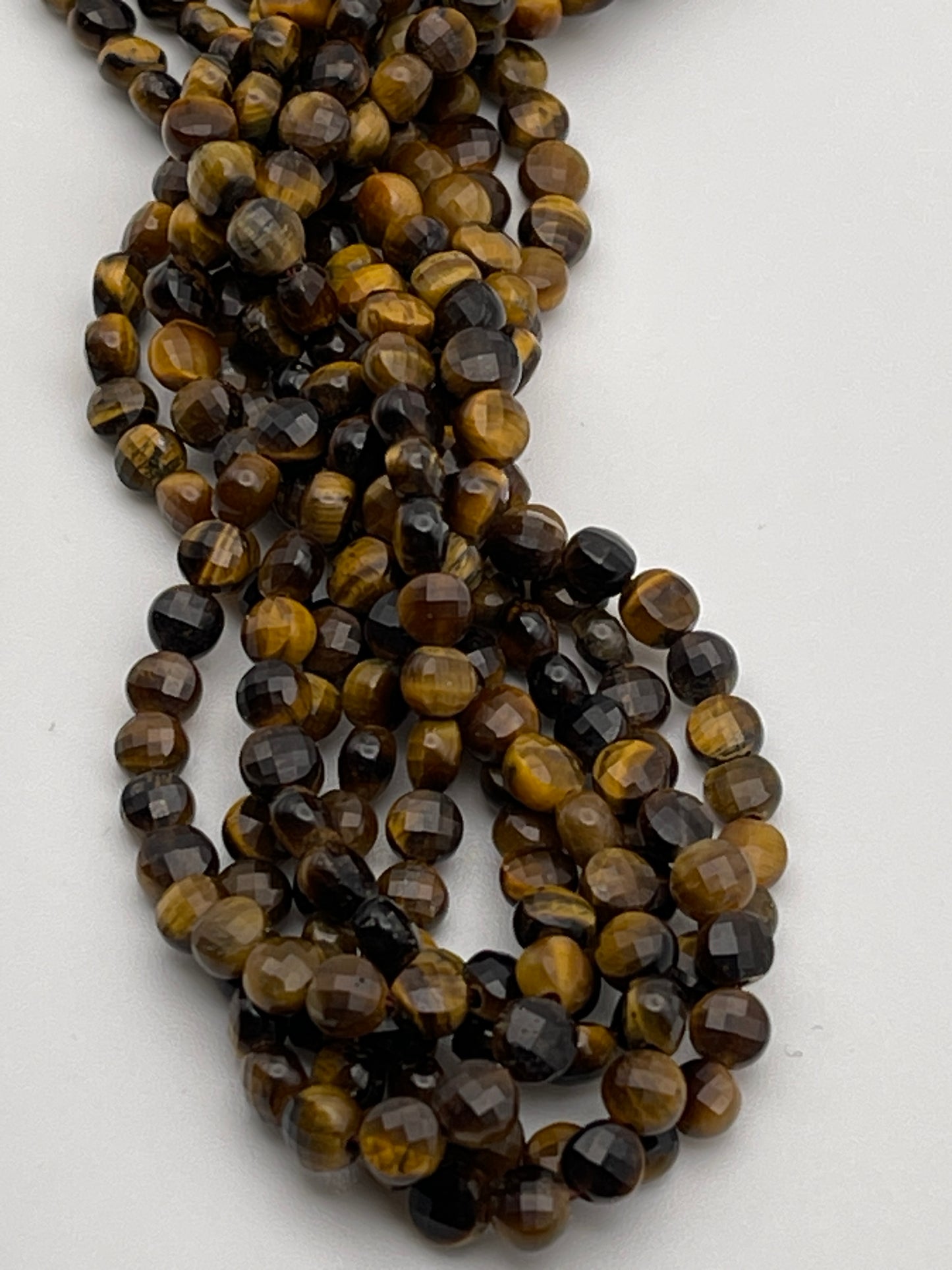 A close-up image of 5mm faceted coin tiger's eye beads. The beads are round and have a glossy finish, showcasing shades of golden brown, black, and dark yellow. The strands are intertwined, creating a textured and intertwined appearance.