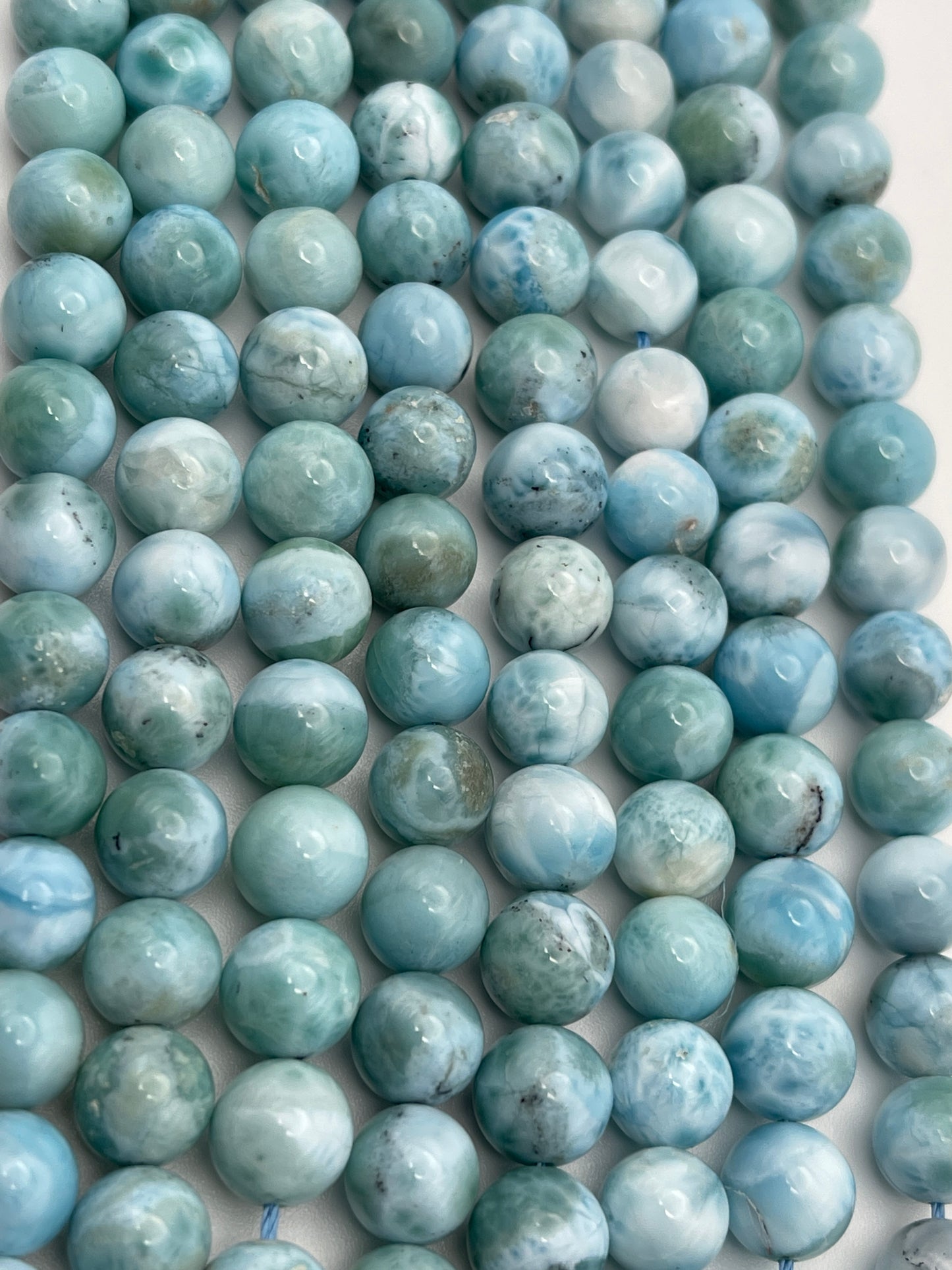 The image shows multiple strands of 8mm Round Larimar beads in various shades of blue and green. The beads are closely packed together and display a marbled pattern with subtle variations in color and texture, ranging from light to dark hues.