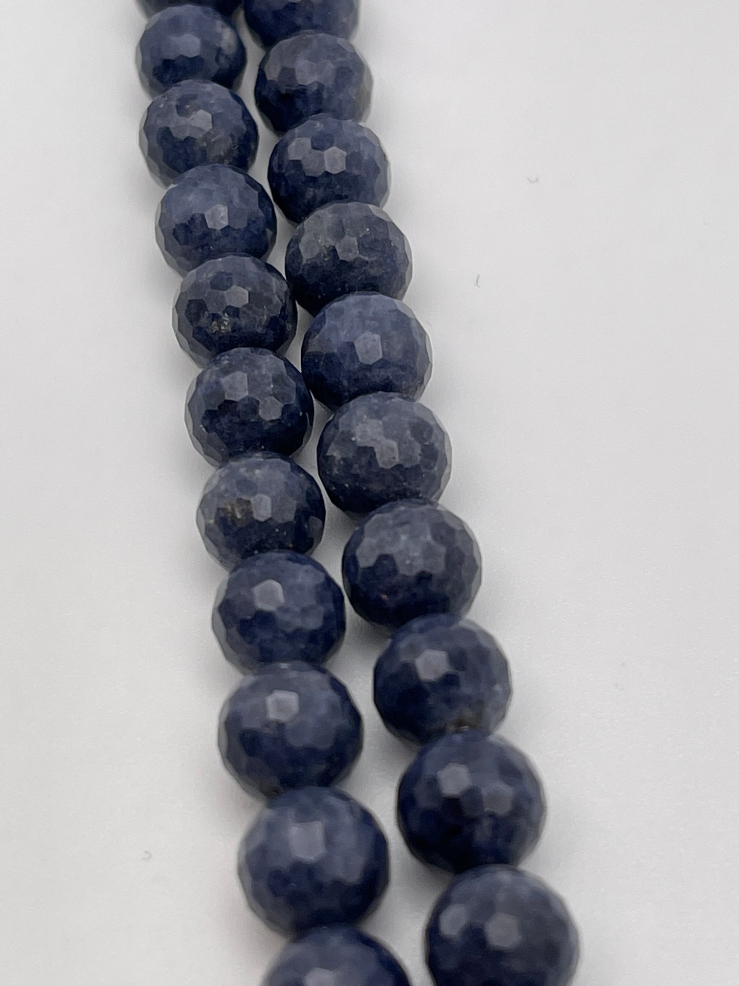 A close-up image of a string of 8mm Fac Sapphire Rnd Blue beads. The dark blue, faceted round beads are arranged in a neat, parallel line, showcasing their smooth, polished texture and a subtle shine that reflects the light. The plain white background highlights the beads' vibrant color and intricate facets.