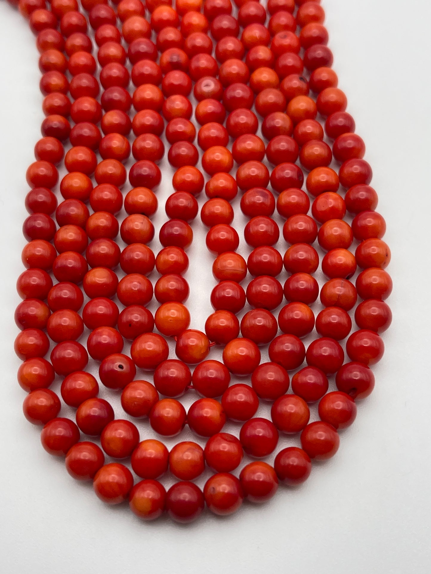 A close-up of multiple strands of 6mm Round Coral beads. The beads are arranged in neat, parallel rows on a white surface. The shades of red vary slightly, creating a vibrant and visually appealing pattern.