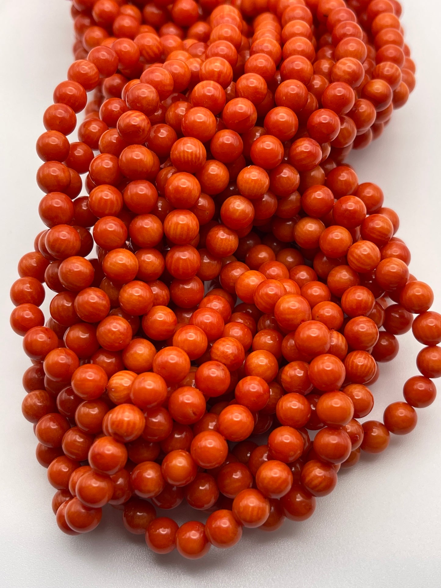 A close-up of several strands of 6mm Round Coral beads, small, glossy, and bright orange, arranged in a loose pile on a white surface. The beads have a smooth texture and uniform color, creating a vibrant and striking visual effect.