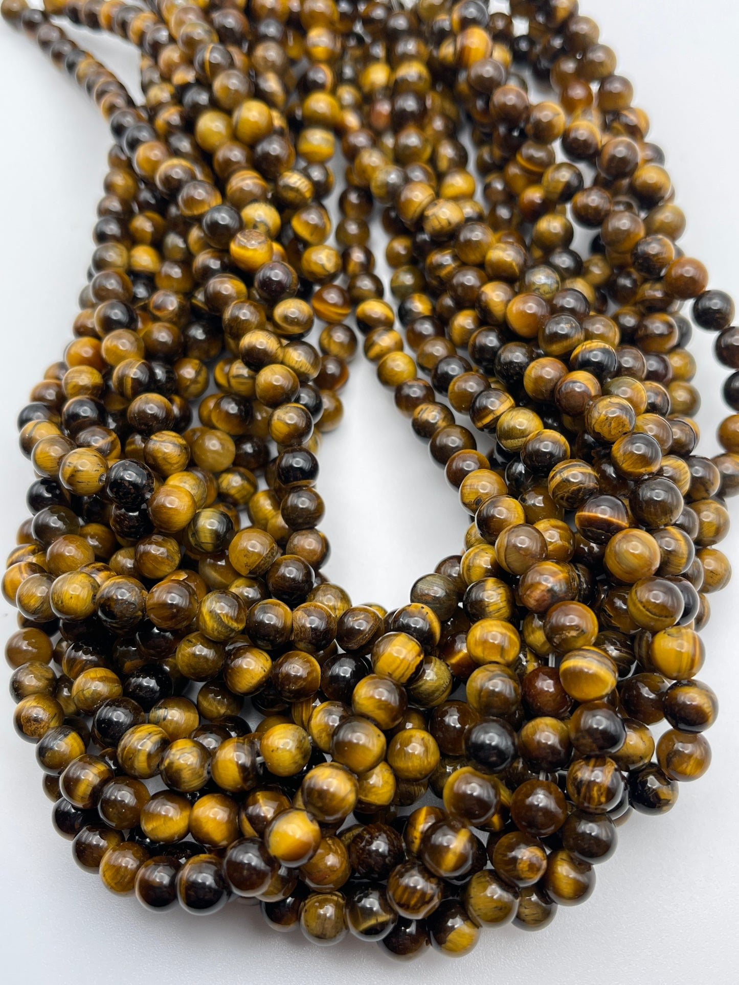 Close-up of multiple strands of 6mm Tiger's Eye Round beads in various shades of brown and yellow, arranged in a layered, intertwined fashion against a light background. The beads have a glossy finish and showcase the characteristic chatoyancy of tiger eye stones.