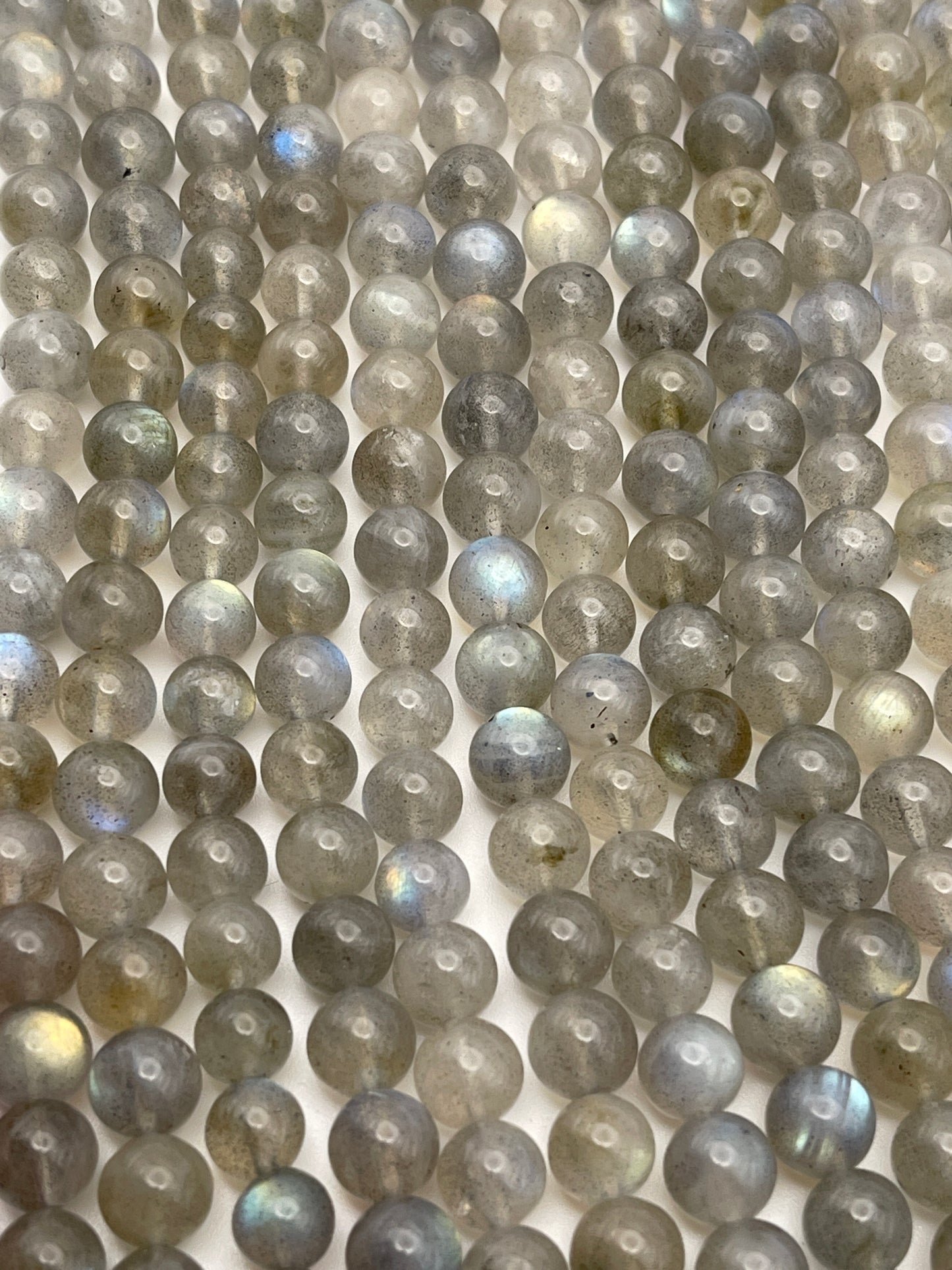 Rows of 7mm Round Labradorite beads with a grey base color and subtle iridescent flashes of blue and green. The semi-translucent beads are evenly spaced and have small holes in their centers, making them ideal for stringing into jewelry or other craft projects.