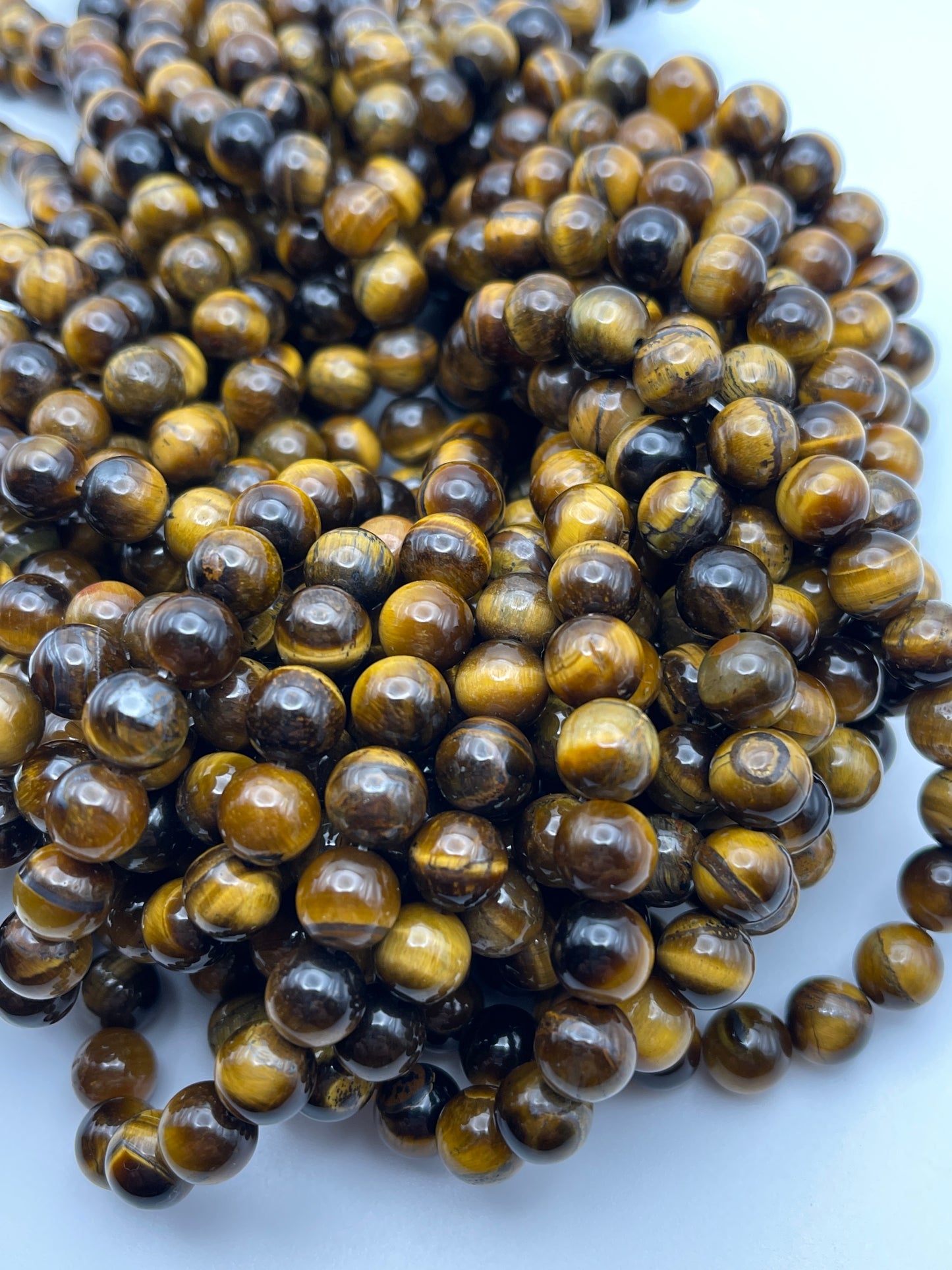 A close-up image of multiple strands of 8mm Round Tiger's Eye beads. The round, polished beads exhibit shades of brown and gold, showcasing a striking chatoyant (cat's-eye) effect. They are arranged closely together in a dense, overlapping manner.