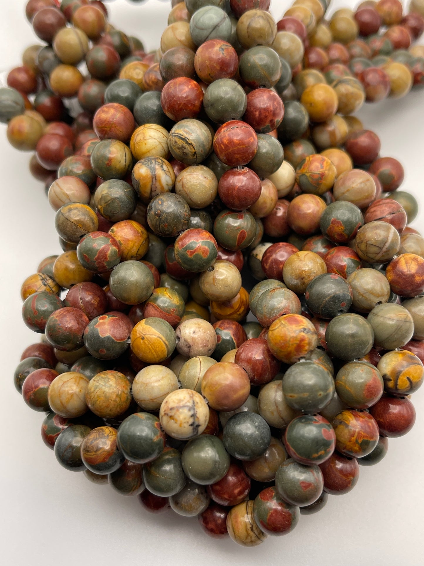 Close-up of several strands of 8mm Red Creek Round Jasper beads. The polished gemstone beads exhibit a variety of colors, including green, brown, red, and yellow, each adorned with unique, intricate patterns and markings. The strands are intricately intertwined and piled together.