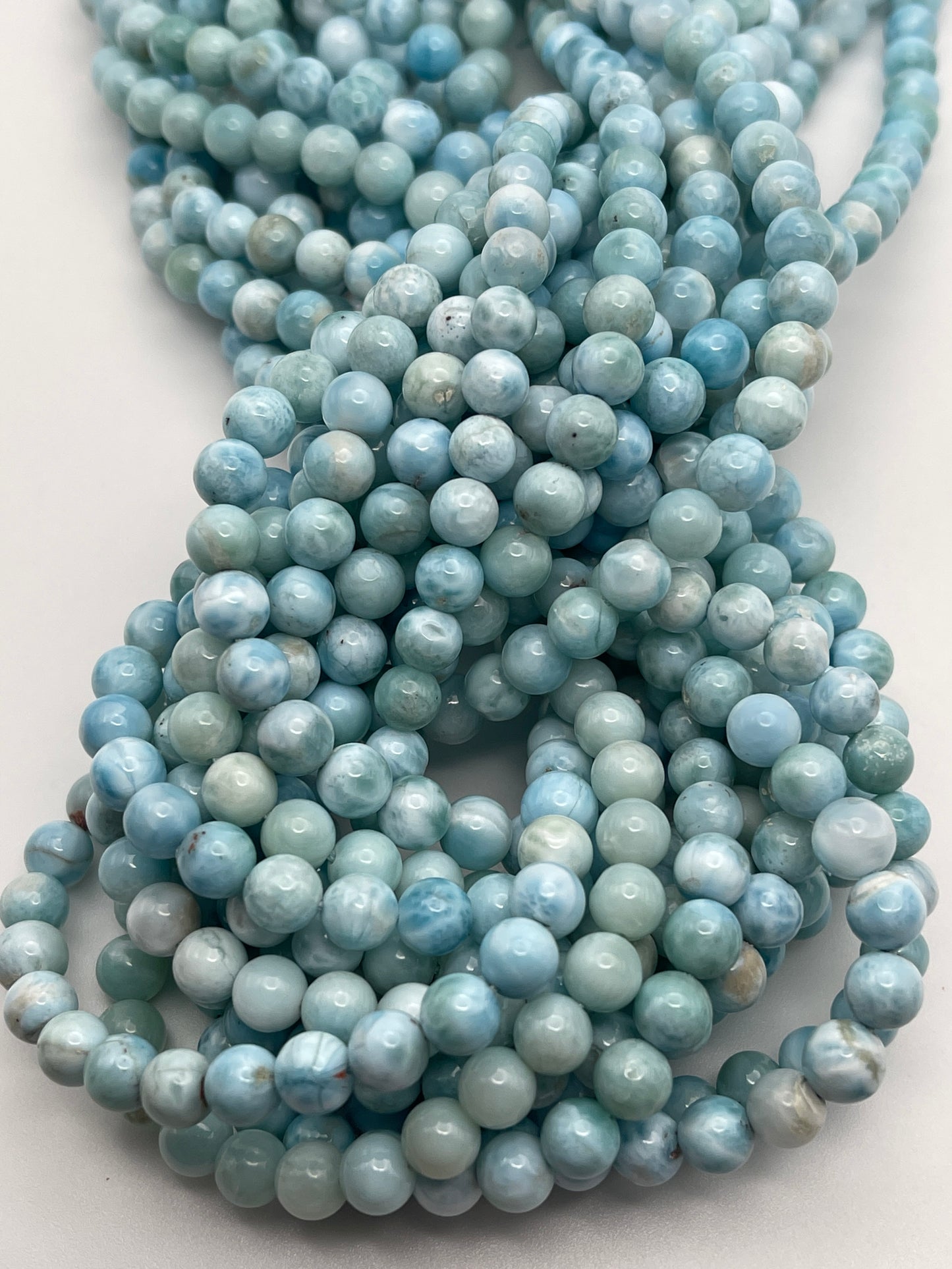 A close-up image of multiple strands of 6mm Round Larimer beads with a smooth, glossy finish. The light blue beads are arranged in an intertwined, cascading manner, creating a textured and intricate pattern. The background is a plain, off-white surface.