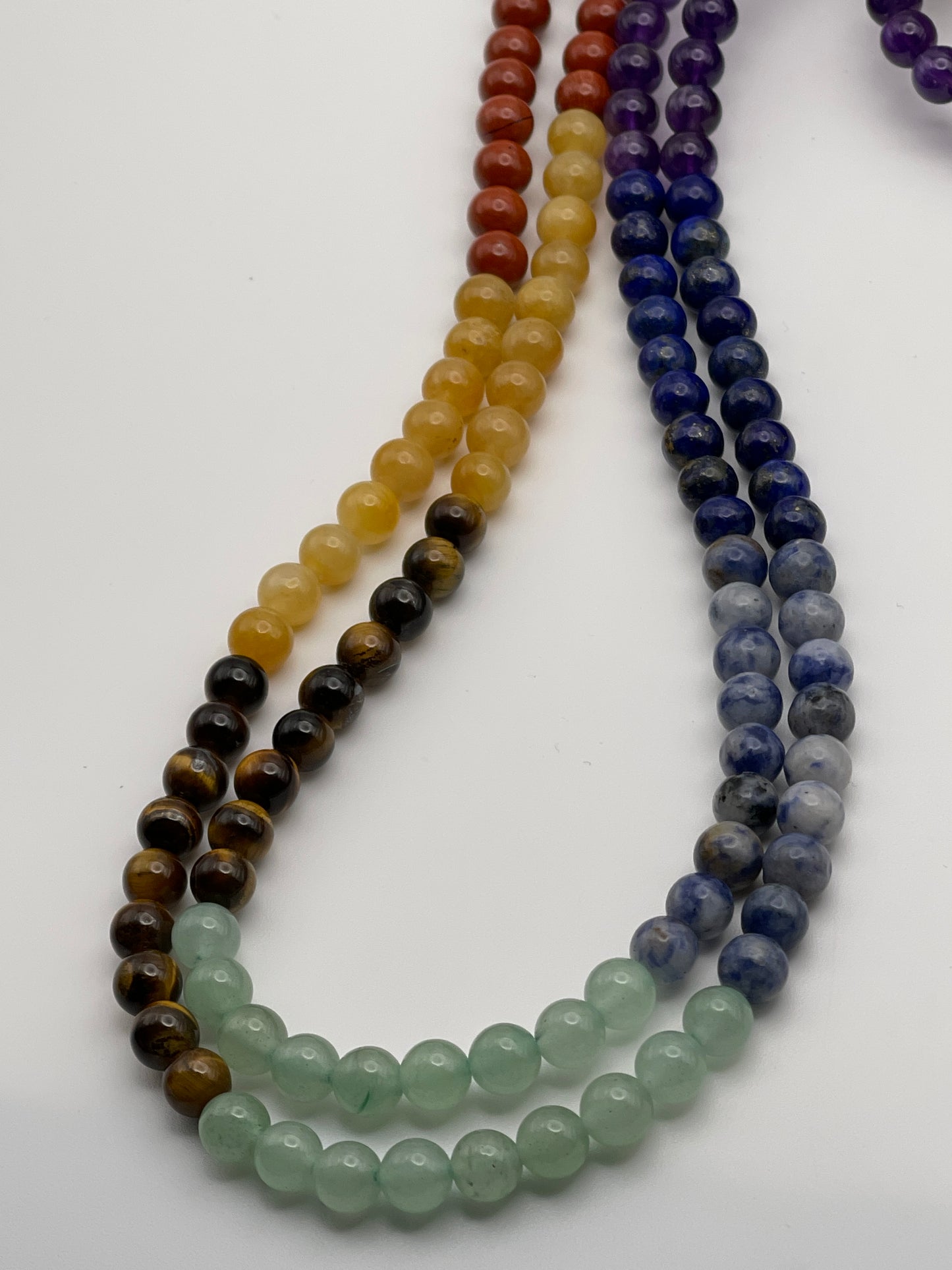 A close-up view of six strands of 6mm Chakra beaded necklaces, each featuring round beads in distinct colors: black, brown, yellow, green, light blue, dark blue, and purple. The necklaces are arranged side by side in a diagonal direction.