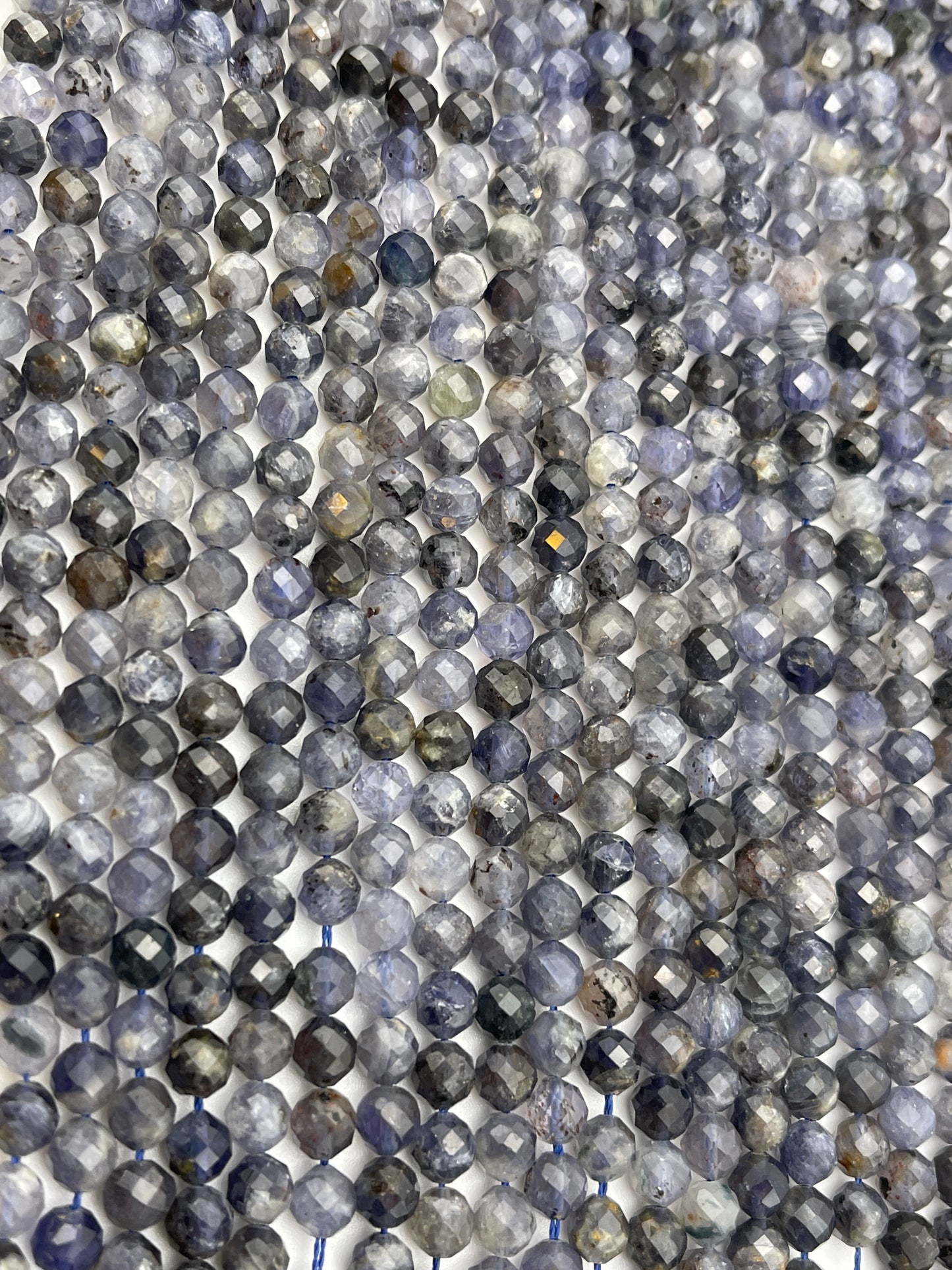 Close-up of numerous strands of 6mm Fac Iolite round beads in various shades of gray, blue, and a hint of brown. The densely packed beads catch the light, creating a shimmering effect across the surface.