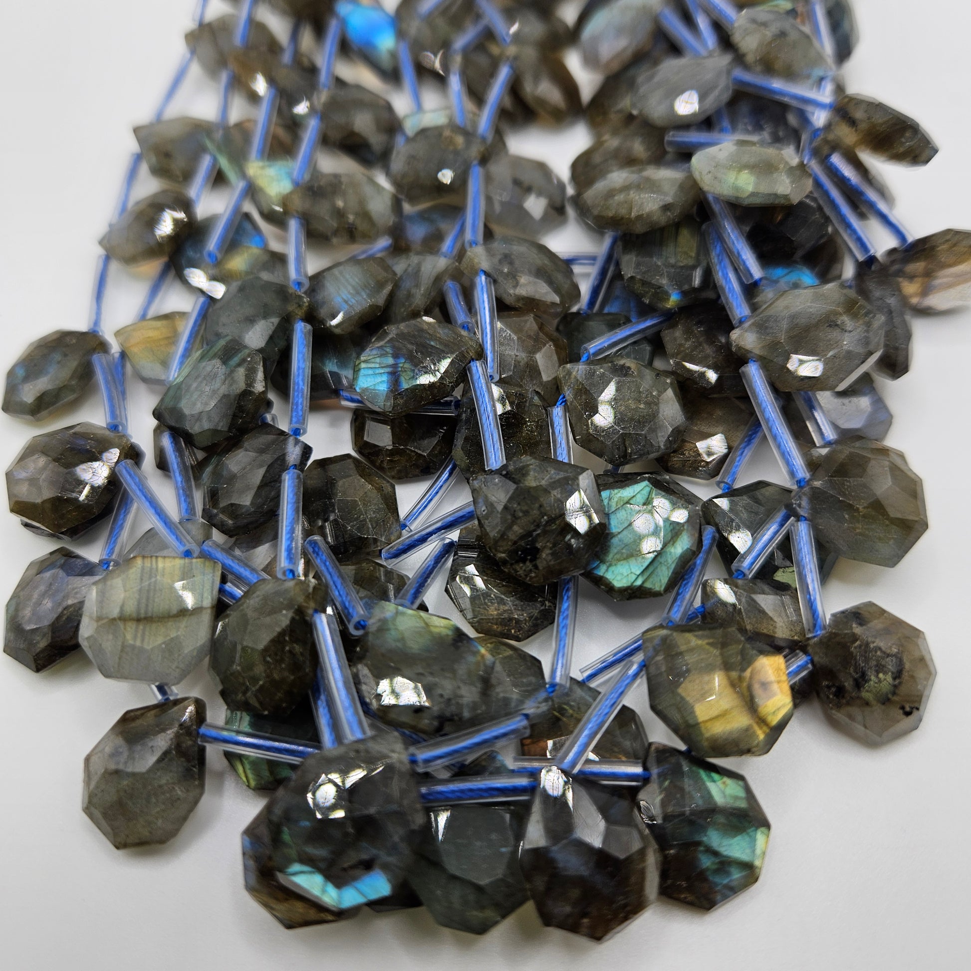 A close-up image features the 13mm Labradorite Faceted Top Drill Bead Strand, displaying multiple strands of irregularly shaped labradorite beads. The white background highlights their visible blue and green flashes, while the blue threads they are strung on accentuate their natural beauty.
