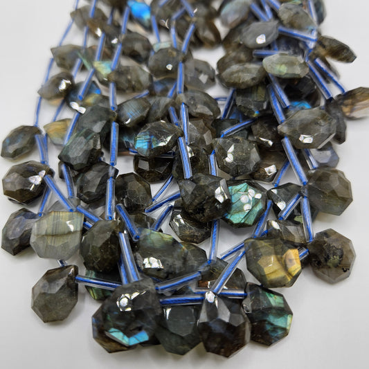 A close-up image features the 13mm Labradorite Faceted Top Drill Bead Strand, displaying multiple strands of irregularly shaped labradorite beads. The white background highlights their visible blue and green flashes, while the blue threads they are strung on accentuate their natural beauty.