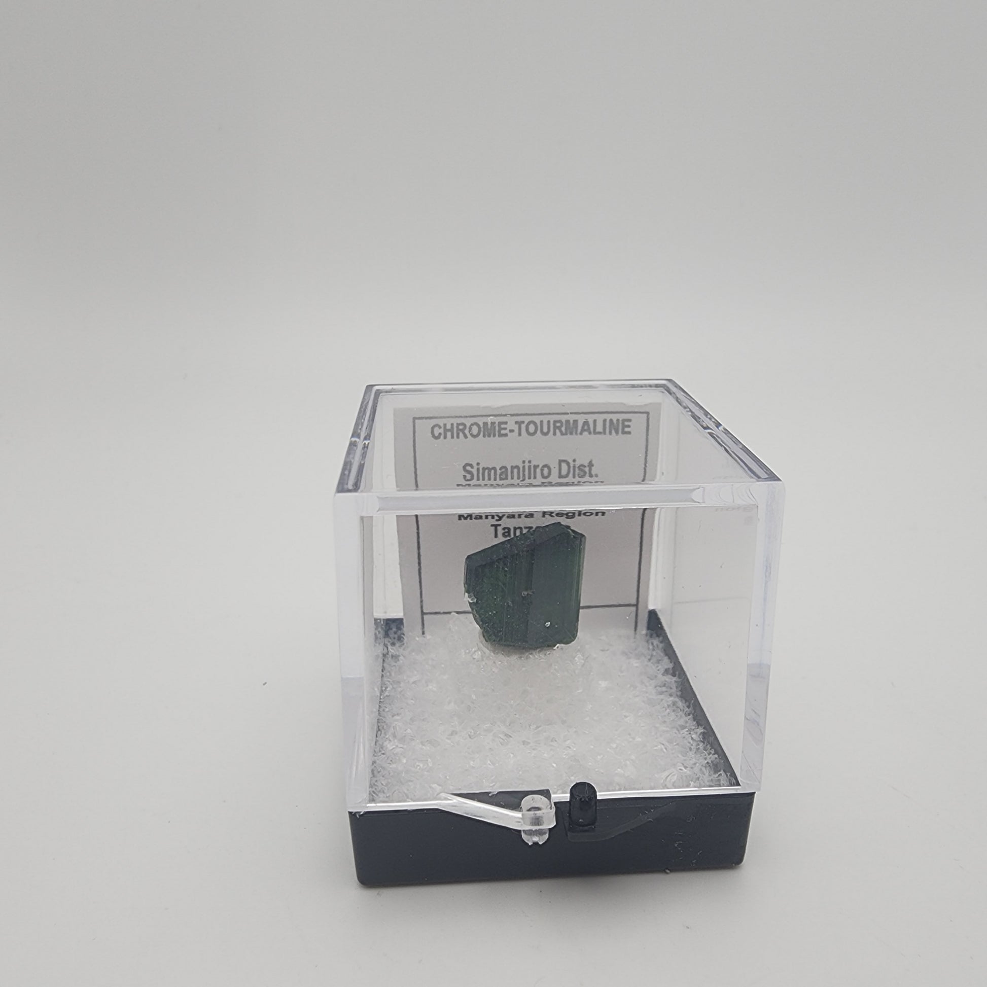 Inside a clear plastic box filled with white cushioning material, a small green Chrome-Tourmaline Specimen is displayed. The label on the box reads "Simanjiro Dist, Manyara Region, Tanzania.