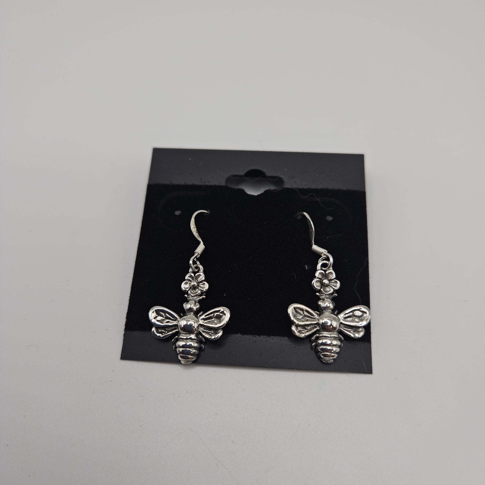 Introducing the SS Bee Earrings, priced at $33.99, featuring a pair of silver earrings intricately designed in the shape of bees with detailed wings and bodies. These stunning earrings are beautifully presented on a black card equipped with hooks for easy display against a neutral gray backdrop.