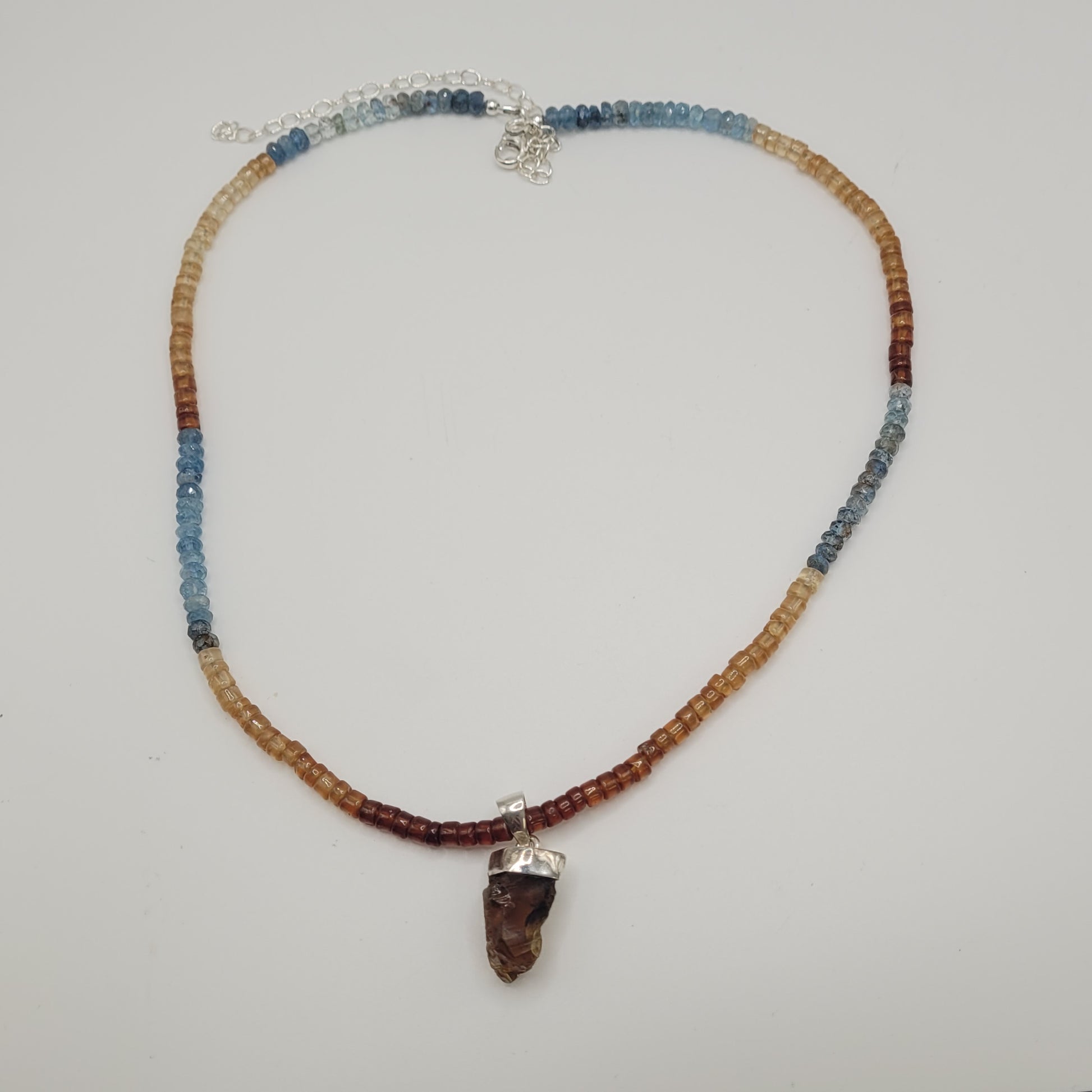 The Congac Quartz, Garnet, and Aquamarine Necklace boasts gradient earth tones, including shades of blue, brown, and tan. It features a prominent dark brown stone pendant set in a silver frame and is secured with a silver lobster clasp with an extension chain. The background is plain white.