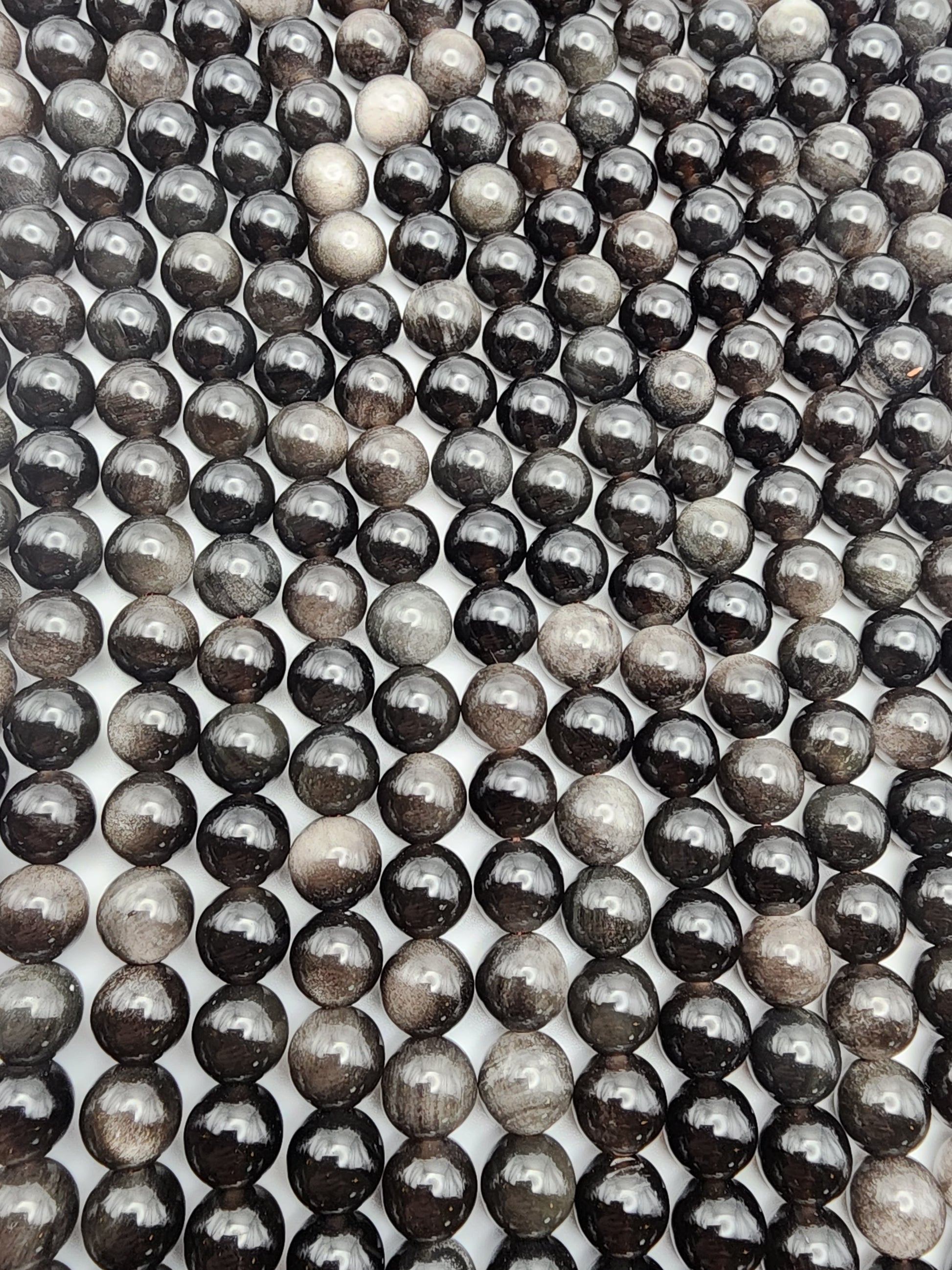 Rows of glossy 8mm Silver Obsidian Round beads, with a black and gray marble-like appearance, are arranged closely together. They create a captivating pattern of reflections and varying shades against a white backdrop.