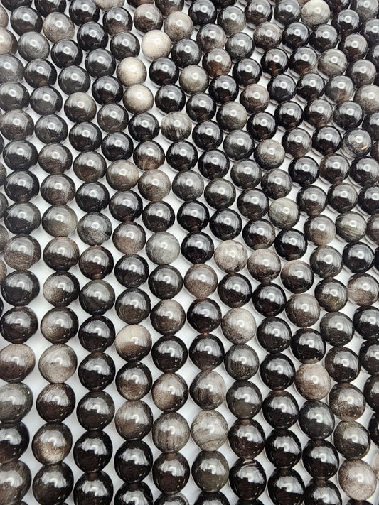 Rows of glossy 8mm Silver Obsidian Round beads, with a black and gray marble-like appearance, are arranged closely together. They create a captivating pattern of reflections and varying shades against a white backdrop.