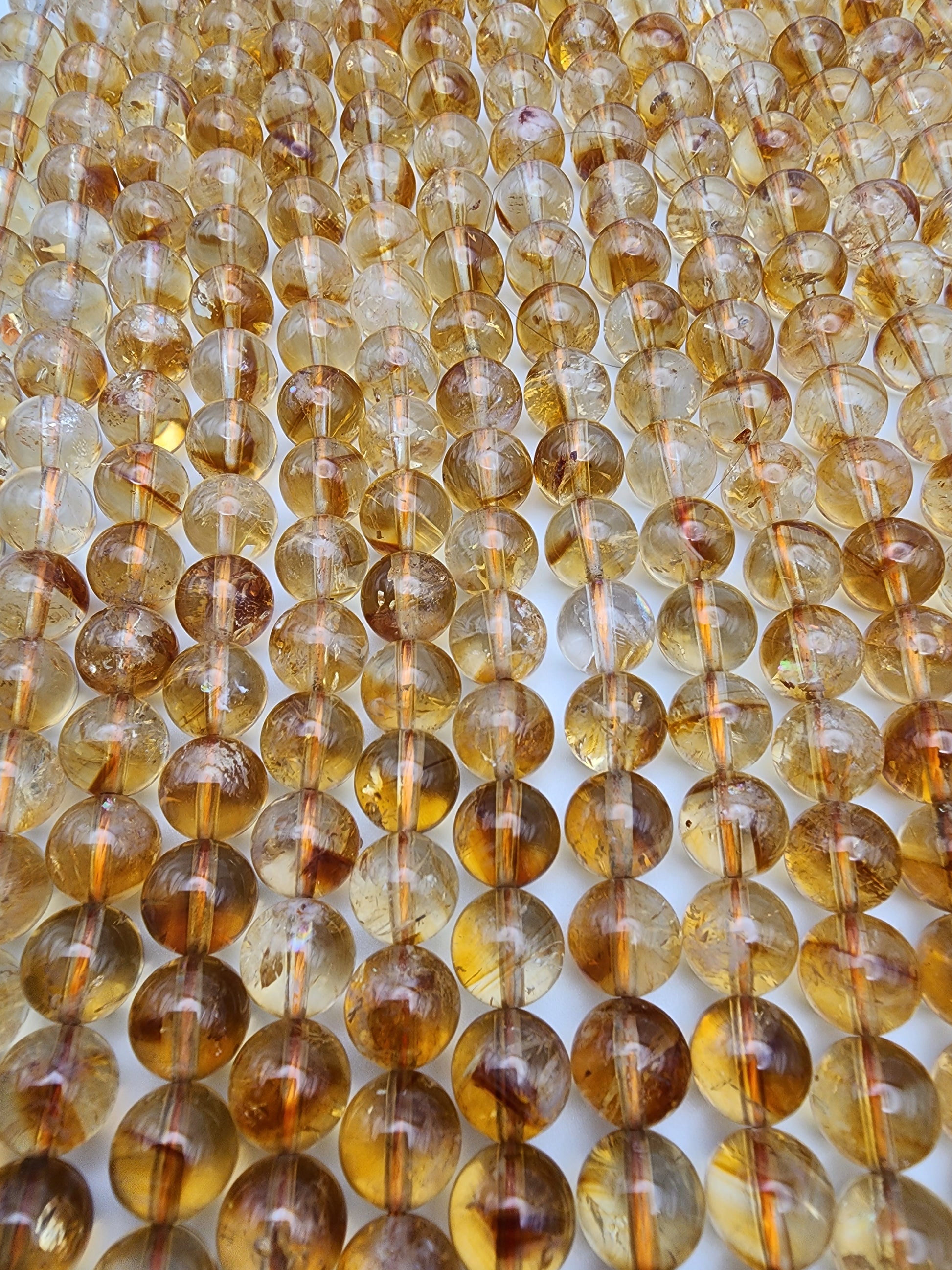 Close-up of several rows of 8mm Citrine Round beads strung together, arranged in parallel lines. The beads have a warm, honey-like color with various inclusions, creating a textured appearance.