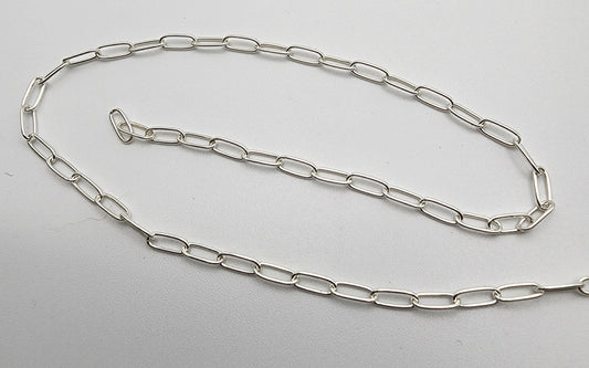 The SS Paper Clip Chain 2.5mm, a delicate silver chain with elongated oval links, is laid out on a white surface. The chain is partially coiled, showcasing its simple design and shine.