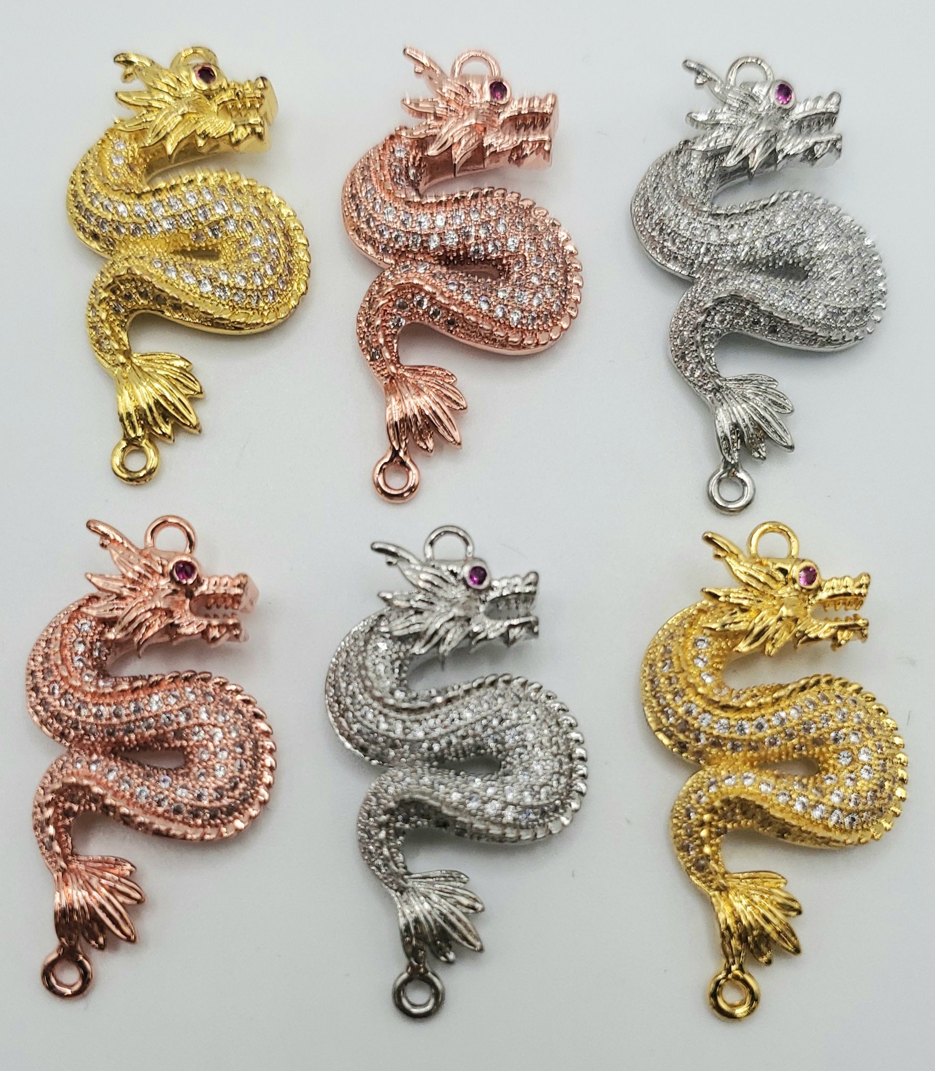 Six Pave Dragon Pendants are arranged in two rows. The top row features pendants in gold, rose gold, and silver. The bottom row consists of rose gold, silver, and gold pendants. Each dragon is encrusted with small crystals and includes a loop for attaching.