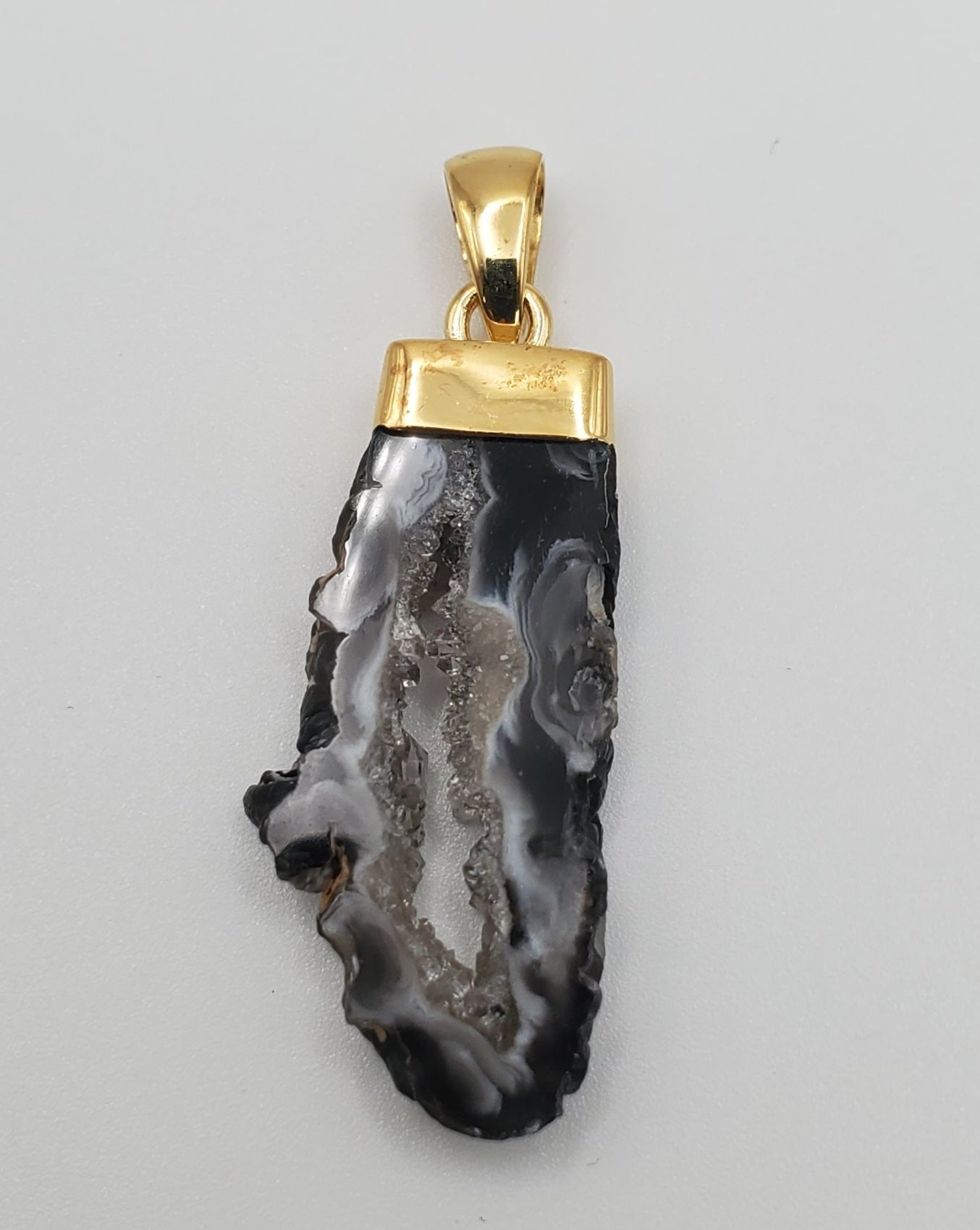 The product description emphasizes the captivating GP Oco Geode Rough Mineral w/ Cap Pendant, featuring a sliced agate stone that displays enchanting swirling gray and black patterns. Encased in a gold-tone cap and bail, the stone's natural rough edges create an organic shape against a light gray background, making it ideal for online sales appeal.
