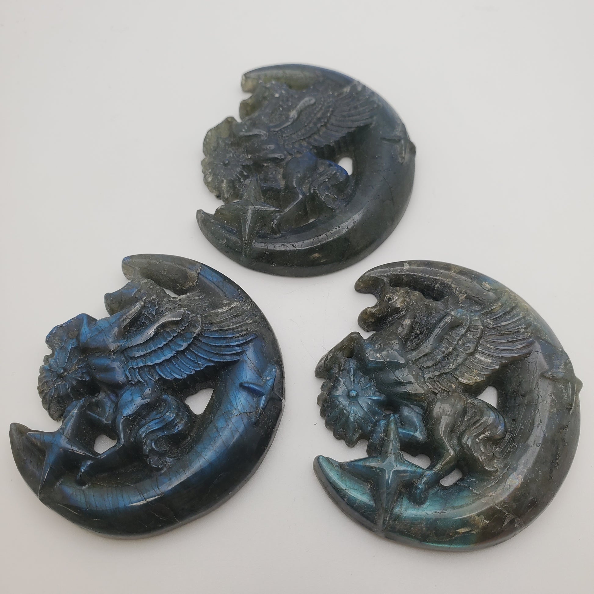 Three "Carved Labradorite Unicorn Moon" sculptures, showcasing winged unicorns perched on crescent moons with a captivating blue iridescent sheen and intricate details, presented against a plain white background.