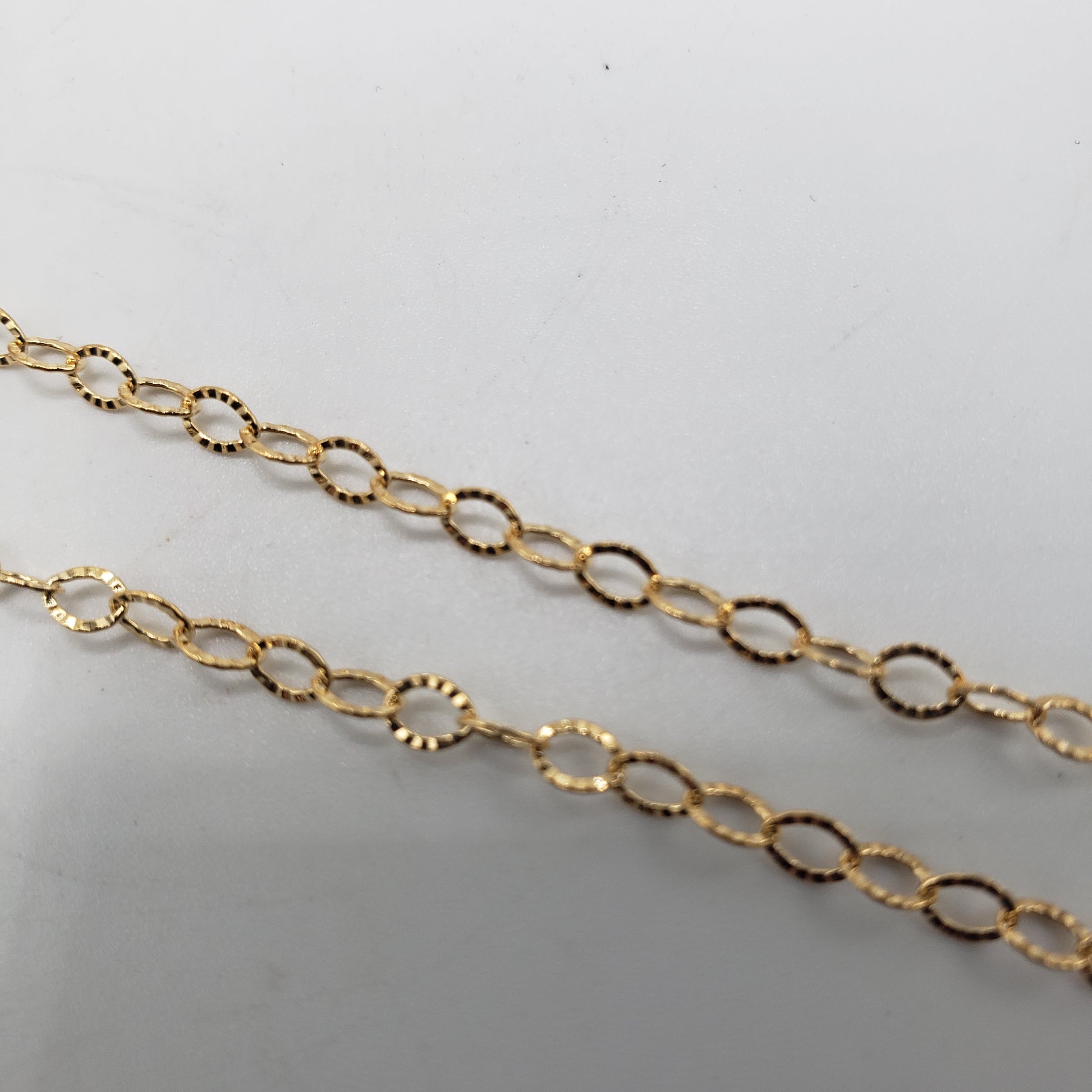The GF Diamond Cut Oval Link Chain is shown against a plain white background, featuring two strands of delicate gold chain with oval links. The close-up view emphasizes the chain's intricate link pattern and glossy finish.