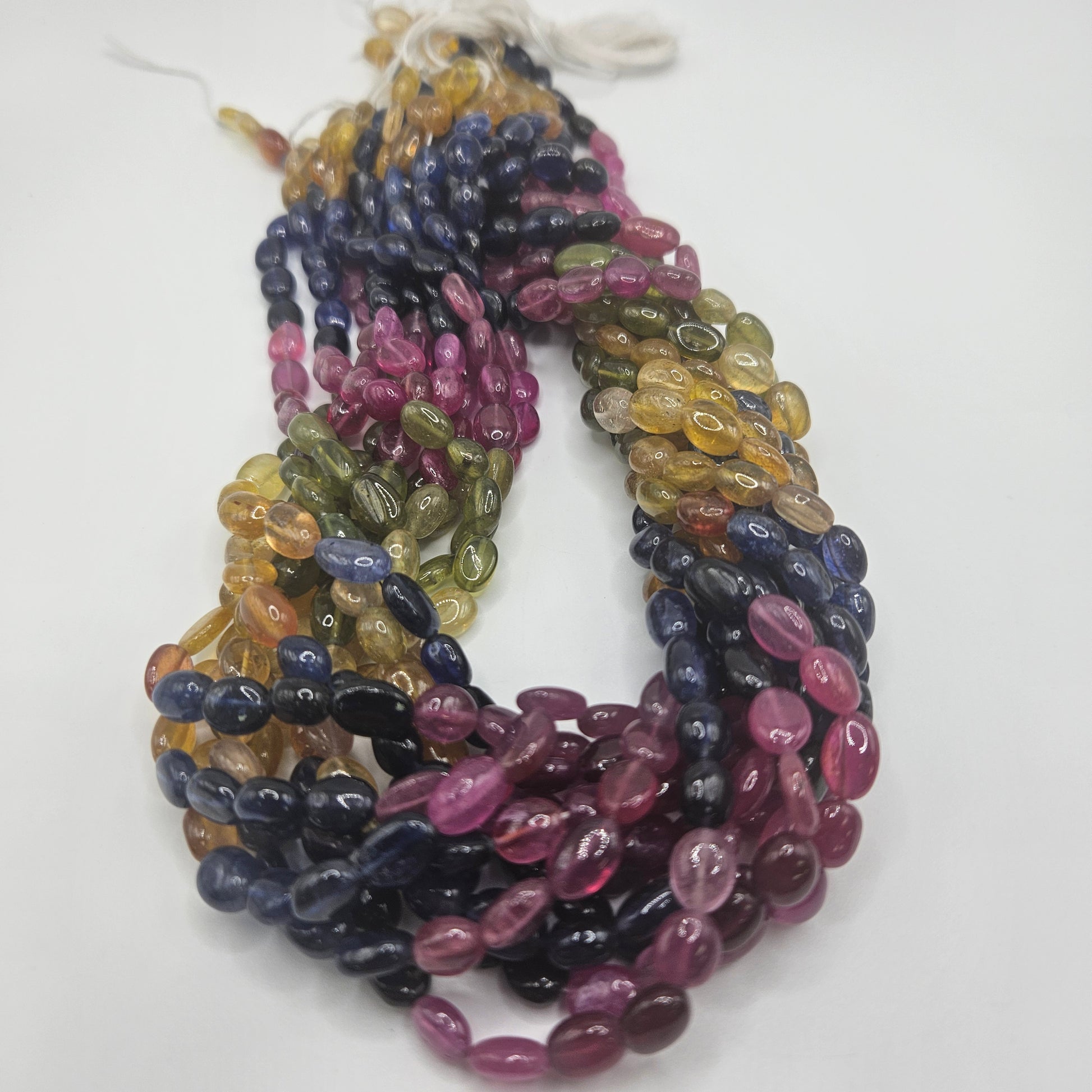 A 6mm Multi Colored Sapphire Oval Bead Strand is displayed on a white surface, showcasing a vibrant array of shades like pink, yellow, green, blue, and dark purple.