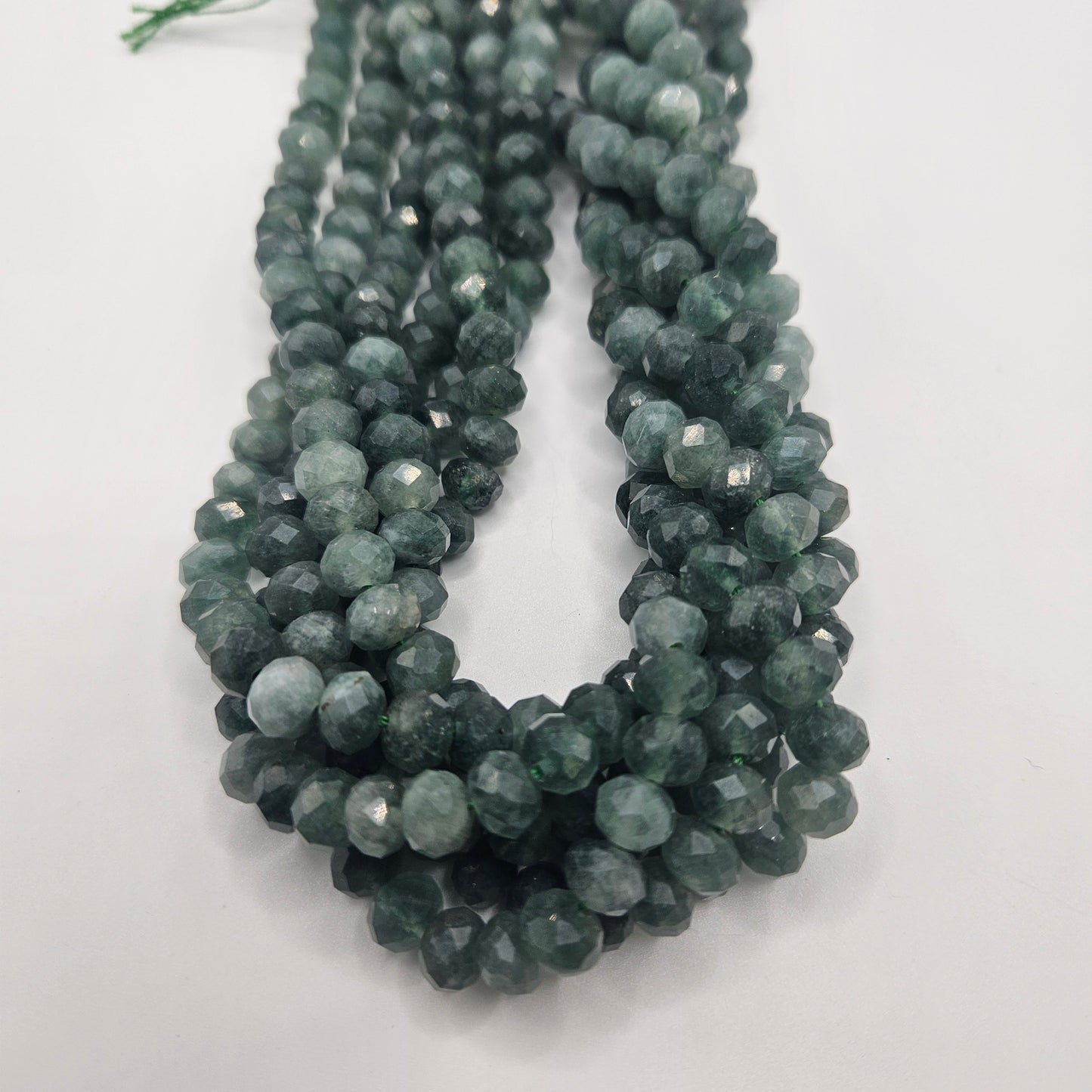 Close-up of multiple strands of the 8mm Green Jade Faceted Rondelle Bead Strand, showcasing their dark green color arranged in a loop on a white background. These beads feature a reflective surface that enhances their texture and vibrancy.