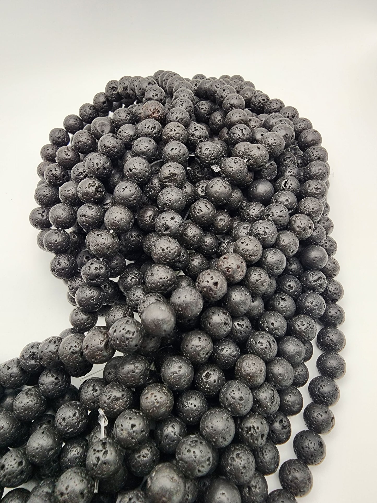 A close-up image showcases a collection of multiple strands of 8mm Lava Rock Rounds spread out in an overlapping manner on a white surface. The black, porous beads have a rough texture and small holes characteristic of volcanic rock. 8mm Lava Rock Round. 
