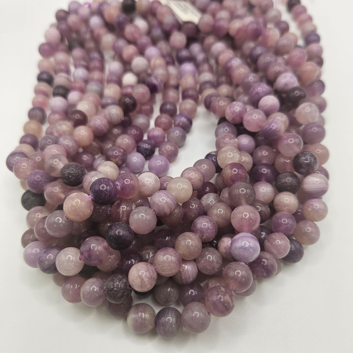 An array of 8mm Purple Fluorite Round Bead Strands, displaying a gradient of shades from light lavender to deep violet, is elegantly looped on a white surface. The enchanting colors hint at the presence of purple fluorite.