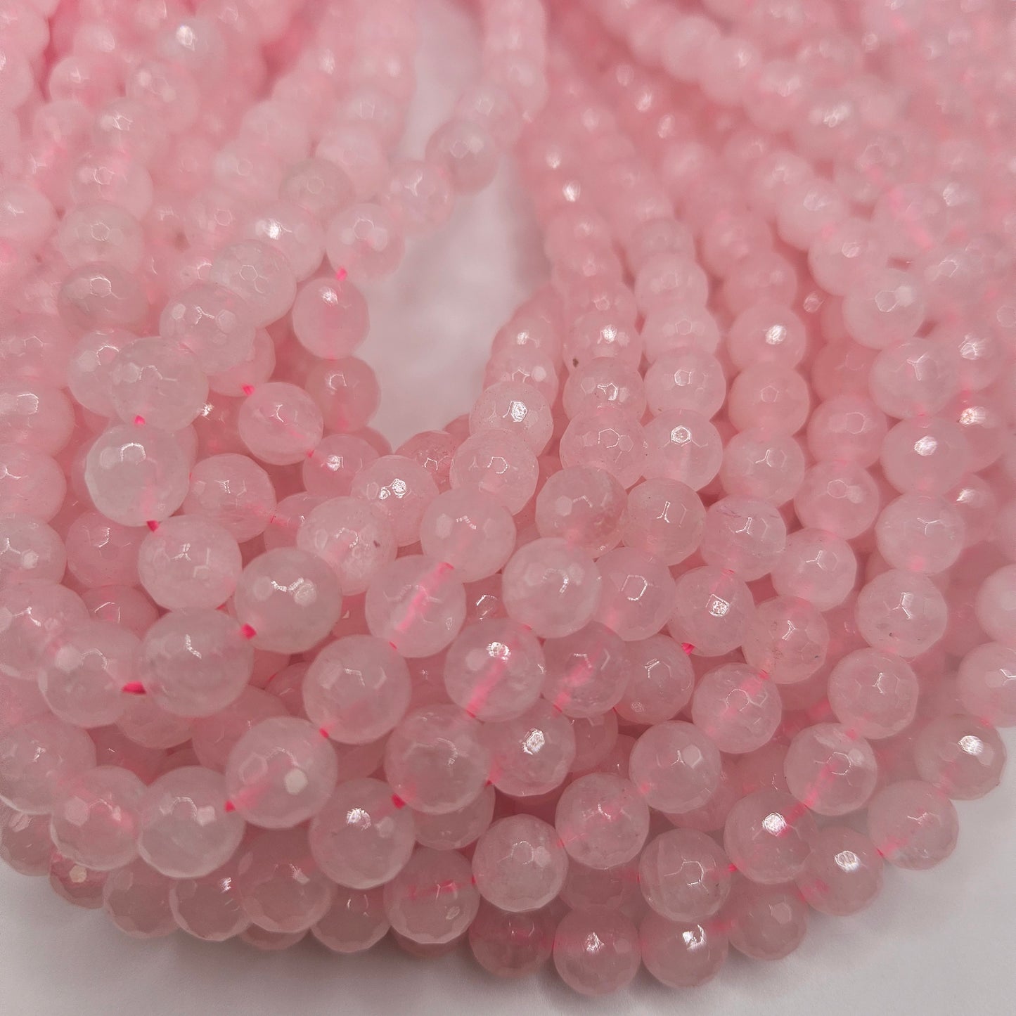 A close-up image features the 8mm Rose Quartz Faceted Bead Strand, with several strands arranged in a loose coil pattern. The faceted beads reflect light, creating a soft, sparkling effect against the white background that enhances their pastel color.
