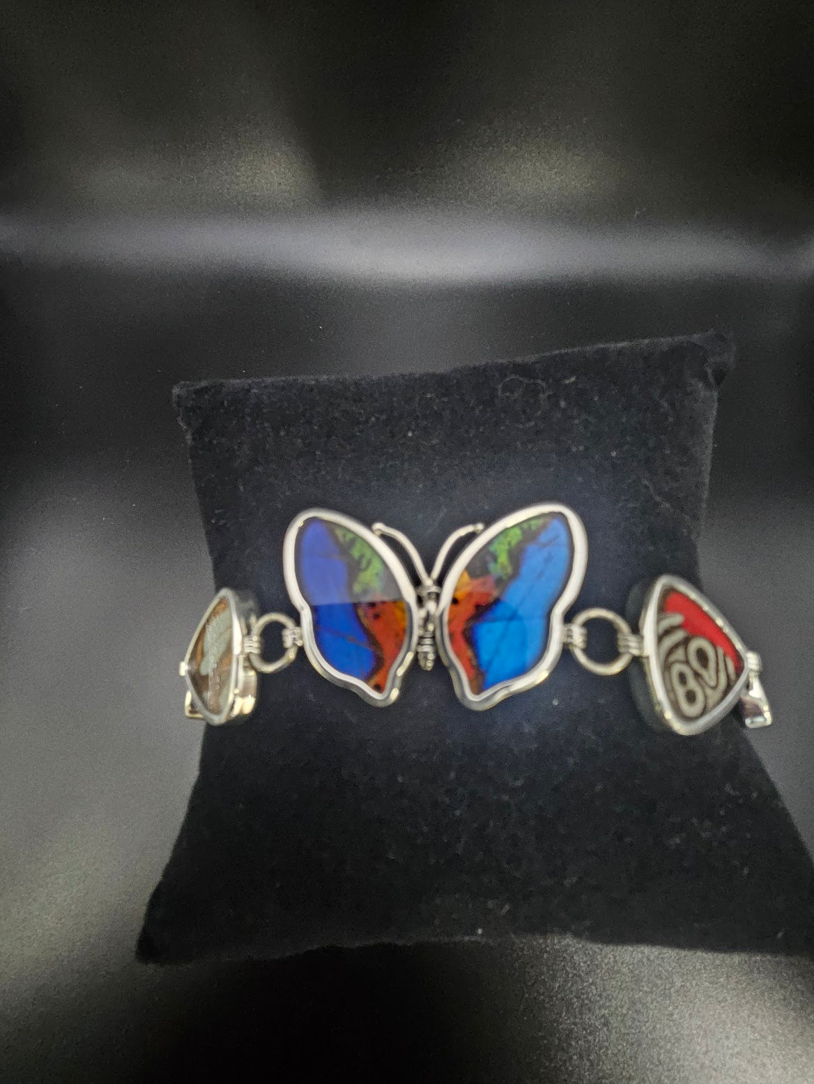 A Sterling Silver 5 Bead Butterfly Bracelet sits elegantly on a black cushion, its wings beautifully adorned with shades of blue, red, and green. This exquisite piece of jewelry captures the vibrant beauty akin to a butterfly sanctuary, featuring intricate decorative elements along its sides.