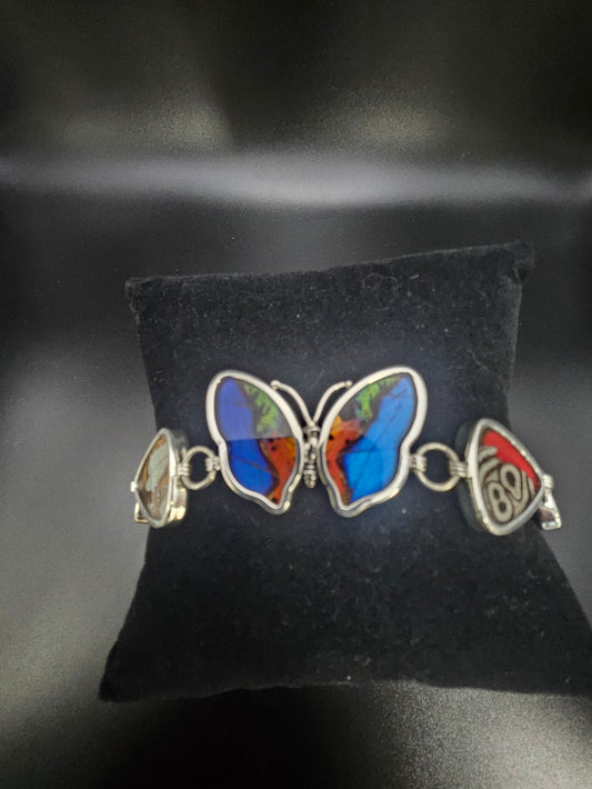 A Sterling Silver 5 Bead Butterfly Bracelet sits elegantly on a black cushion, its wings beautifully adorned with shades of blue, red, and green. This exquisite piece of jewelry captures the vibrant beauty akin to a butterfly sanctuary, featuring intricate decorative elements along its sides.