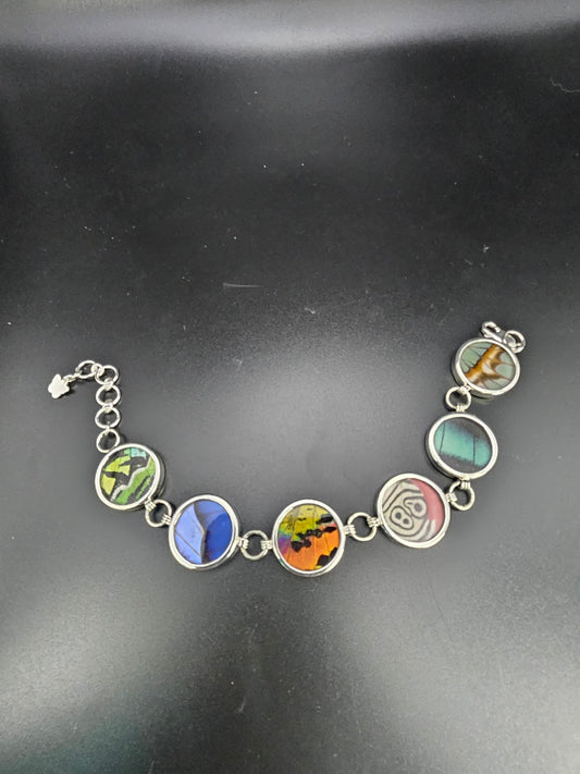 Introducing the Sterling Silver XL 6 Wing Bracelet, a stunning accessory adorned with six round colorful pendants that feature abstract designs set against a black background. Each pendant displays unique and vibrant patterns reminiscent of butterfly jewelry, with shades of green, blue, orange, pink, and beyond.