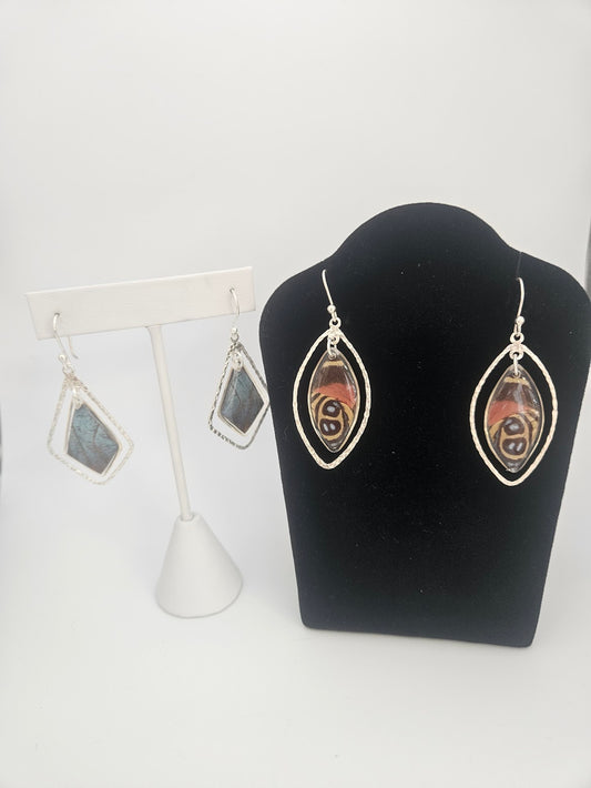 Displayed are two exquisite pairs of earrings: one featuring an abstract pattern on a black stand, and the other, the Sterling Silver Butterfly Ulises Earrings adorned with blue stone pendants reminiscent of delicate butterfly jewelry, showcased on a white stand against a plain background.