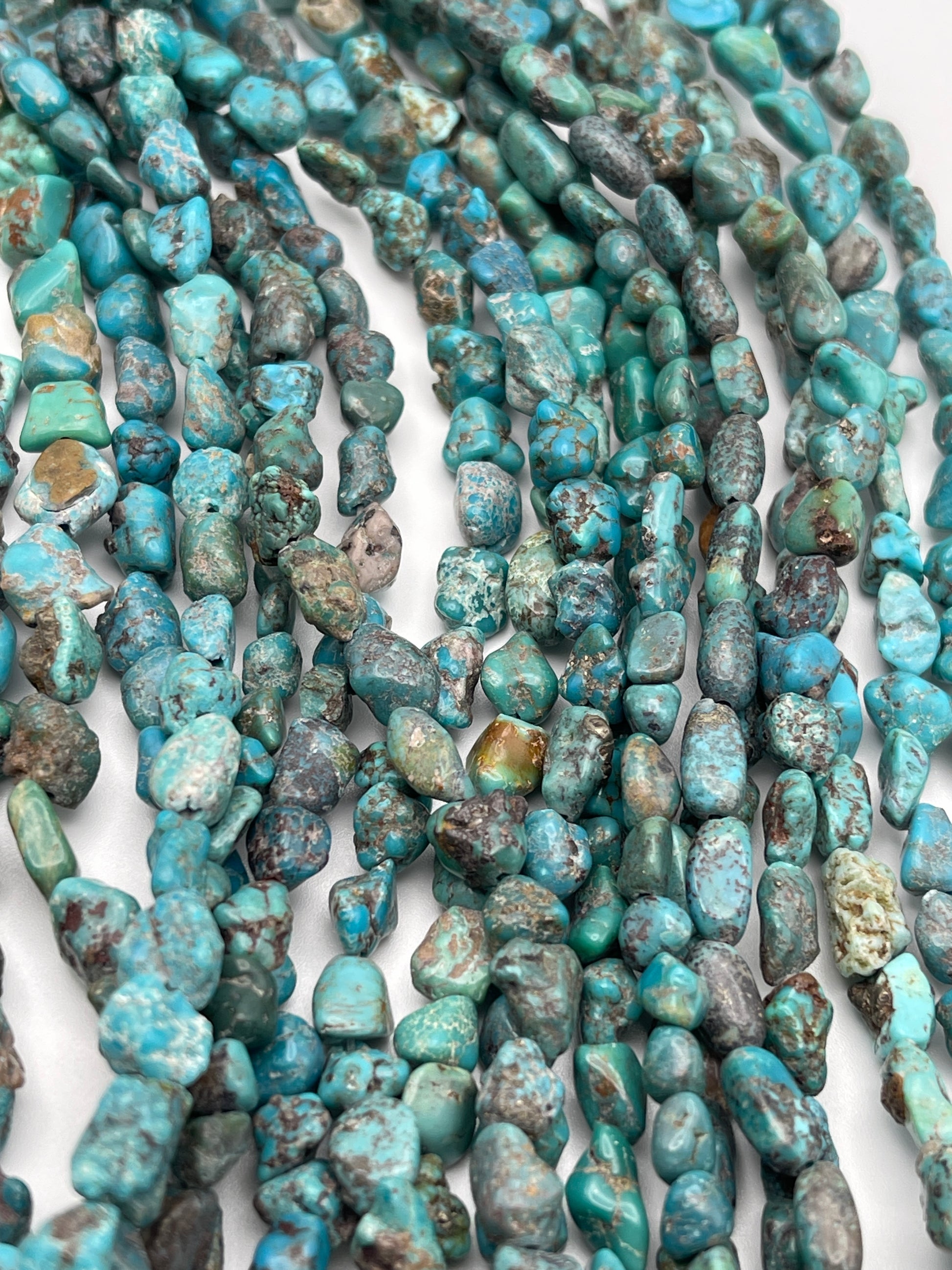 Close-up view of multiple strands of 6-8mm Turquoise Chunk beads, showcasing a variety of blue-green hues with natural veining and textures, set against a plain white background.