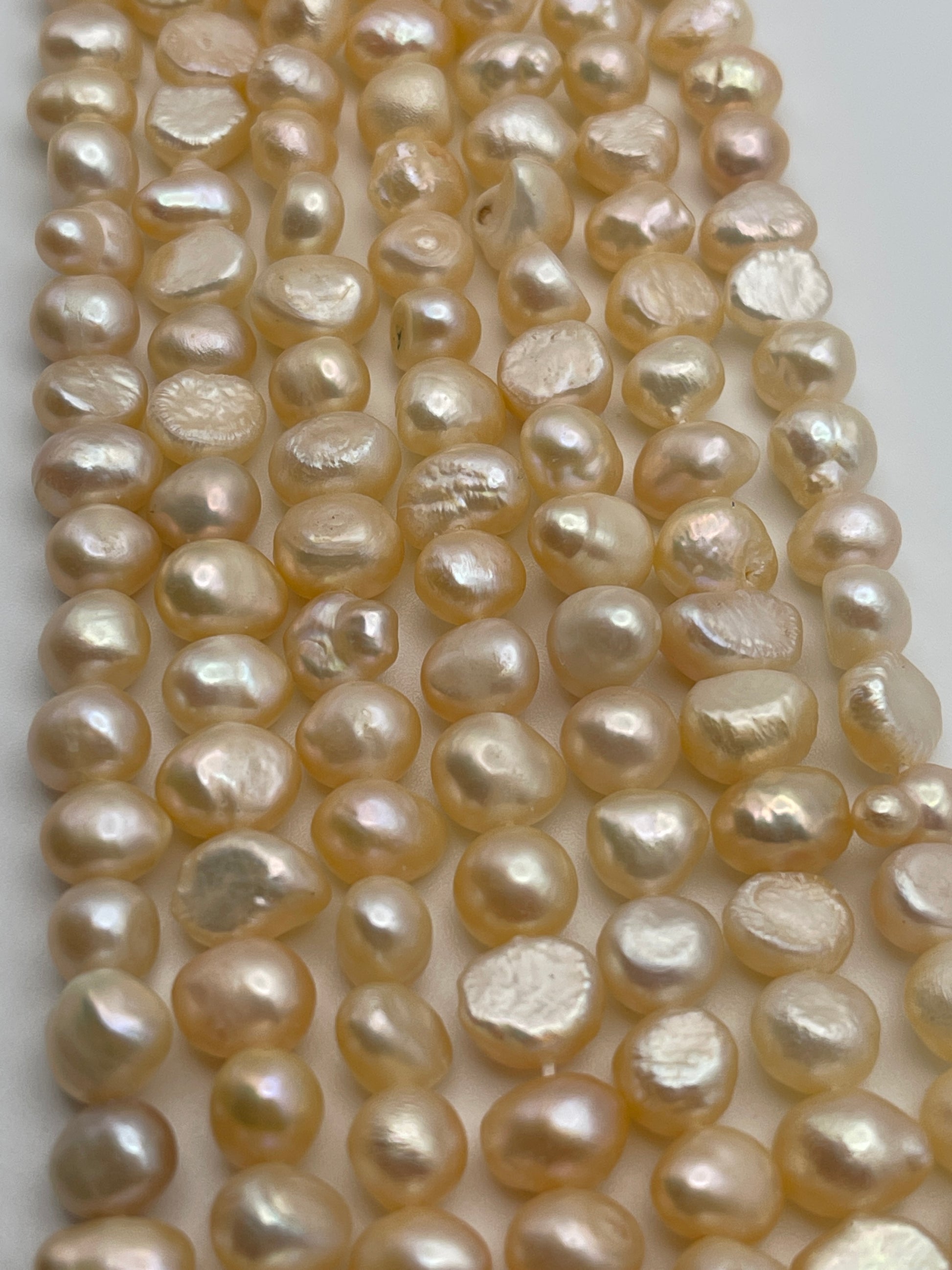 Close-up image of strands of irregularly shaped, peach-colored flat pearls with a subtle luster, laid out in parallel rows on a white surface. The 7-8mm pearls vary slightly in size and shape, giving a natural and organic appearance.