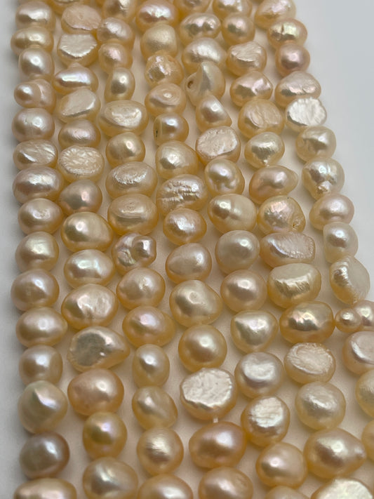 Close-up image of strands of irregularly shaped, peach-colored flat pearls with a subtle luster, laid out in parallel rows on a white surface. The 7-8mm pearls vary slightly in size and shape, giving a natural and organic appearance.